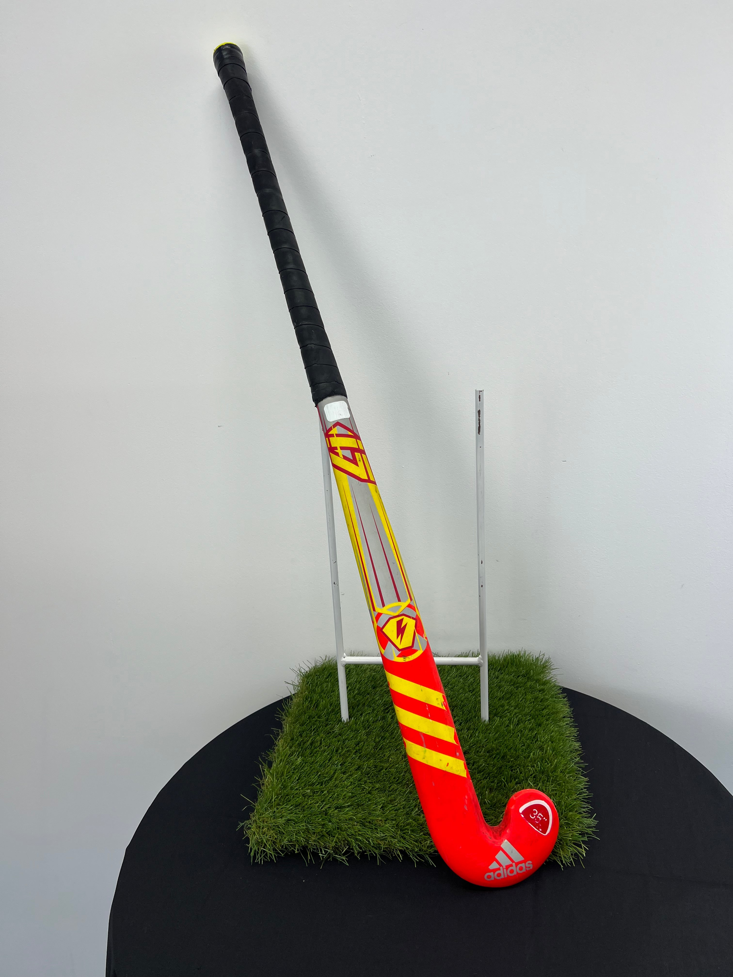Full view of adidas Junior Hockey Stick 35” standing on artificial grass with black wrapped grip and bright red and yellow design