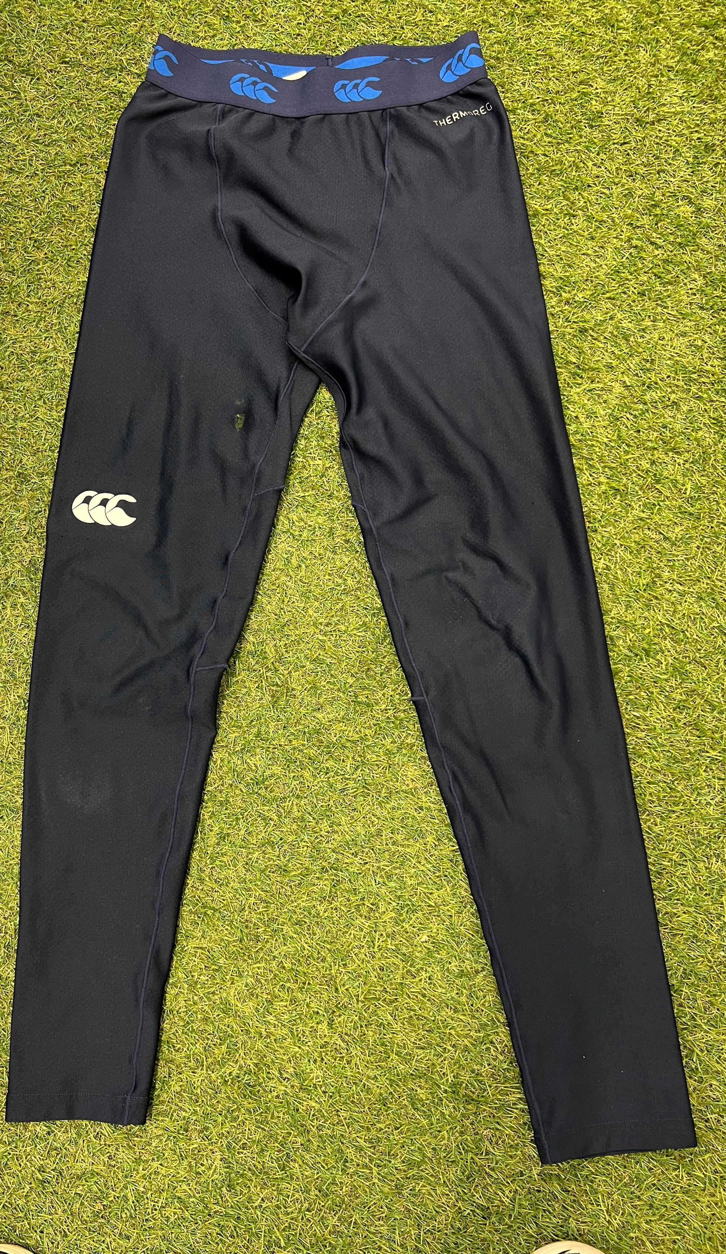 Front view of Canterbury navy base layer leggings laid flat on grass, featuring elastic waistband with blue logos