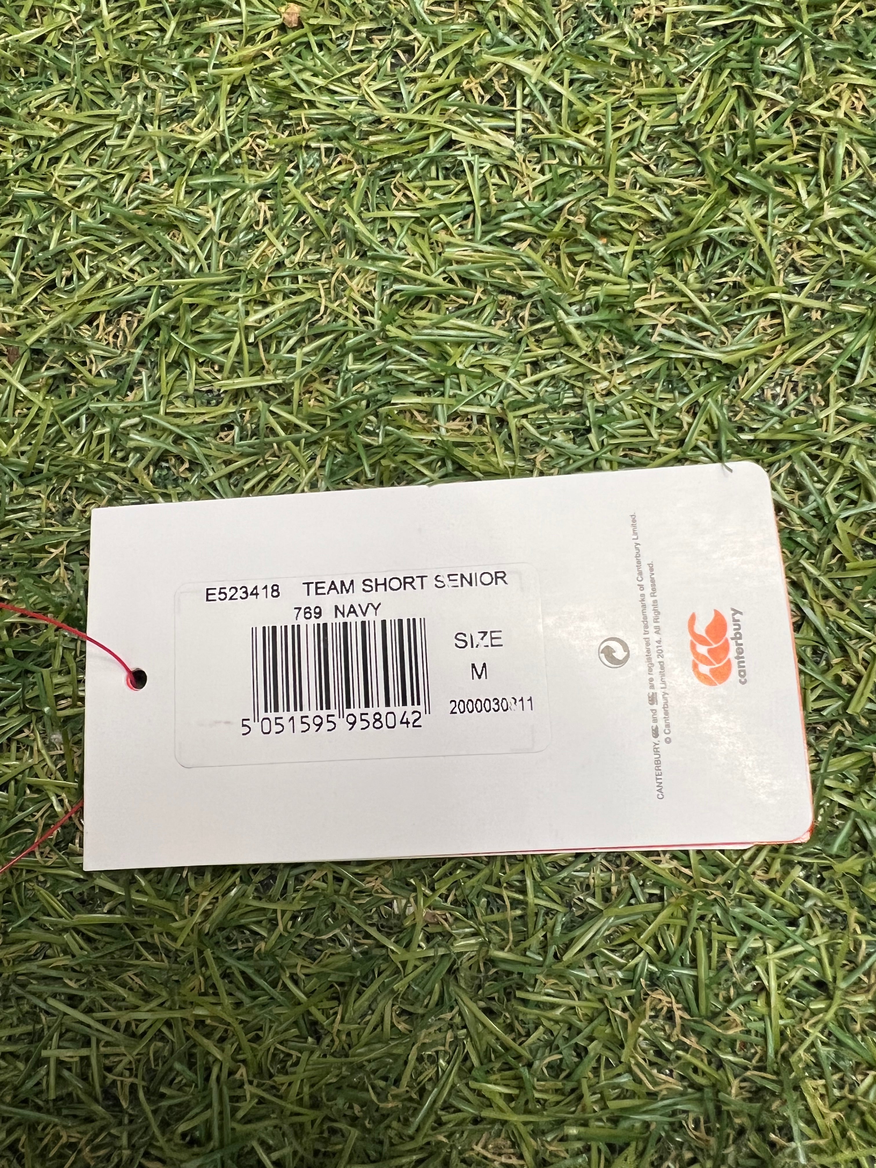 Close-up of product tag with barcode showing 'TEAM SHORT SENIOR', navy color, size M and Canterbury branding