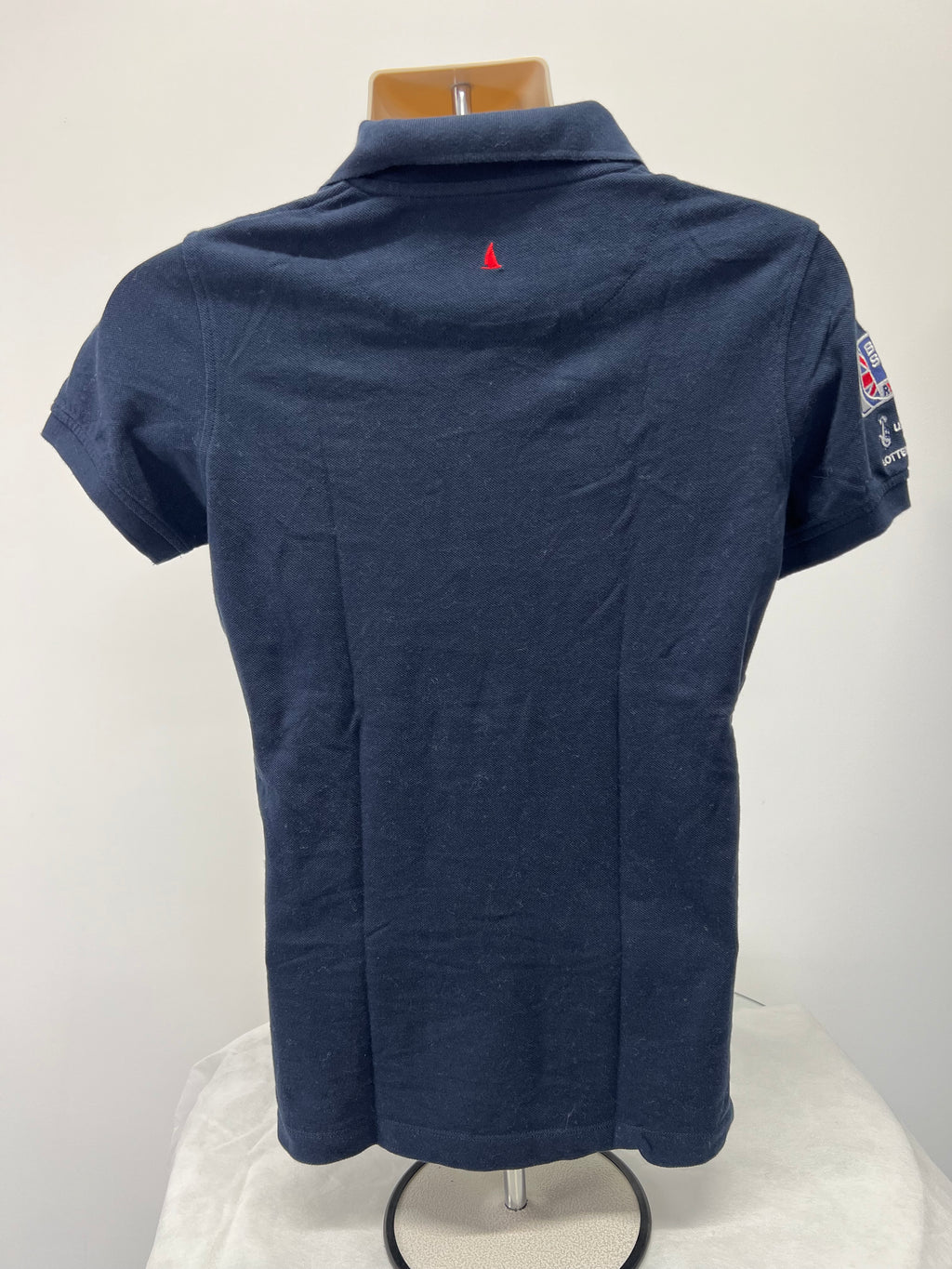 Back view of navy Musto polo shirt showing plain fabric and small red sailboat embroidery near collar