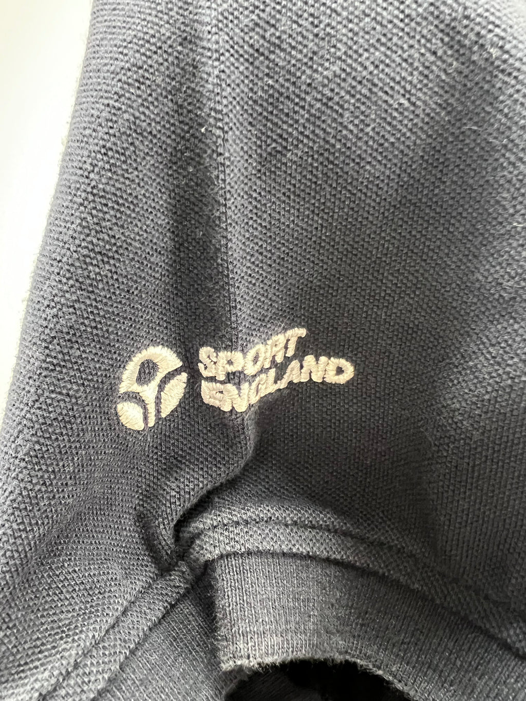 Close-up of navy polo shirt sleeve featuring embroidered Sport England logo in white