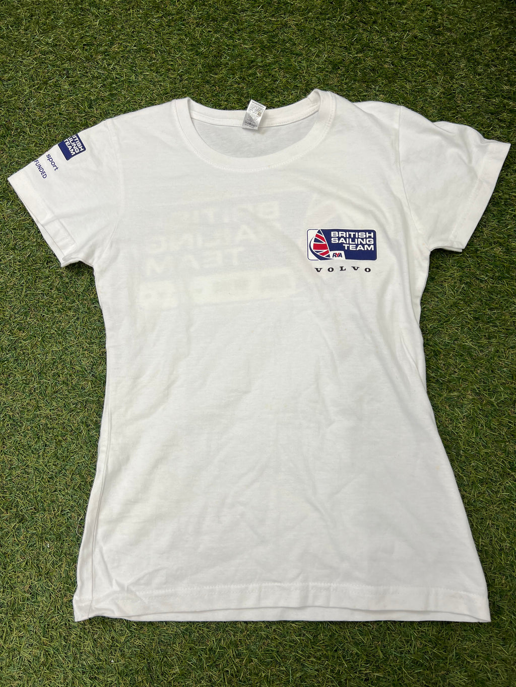 Front view of a white women's British Sailing Team t-shirt with small logo on chest and sleeve branding on grass background