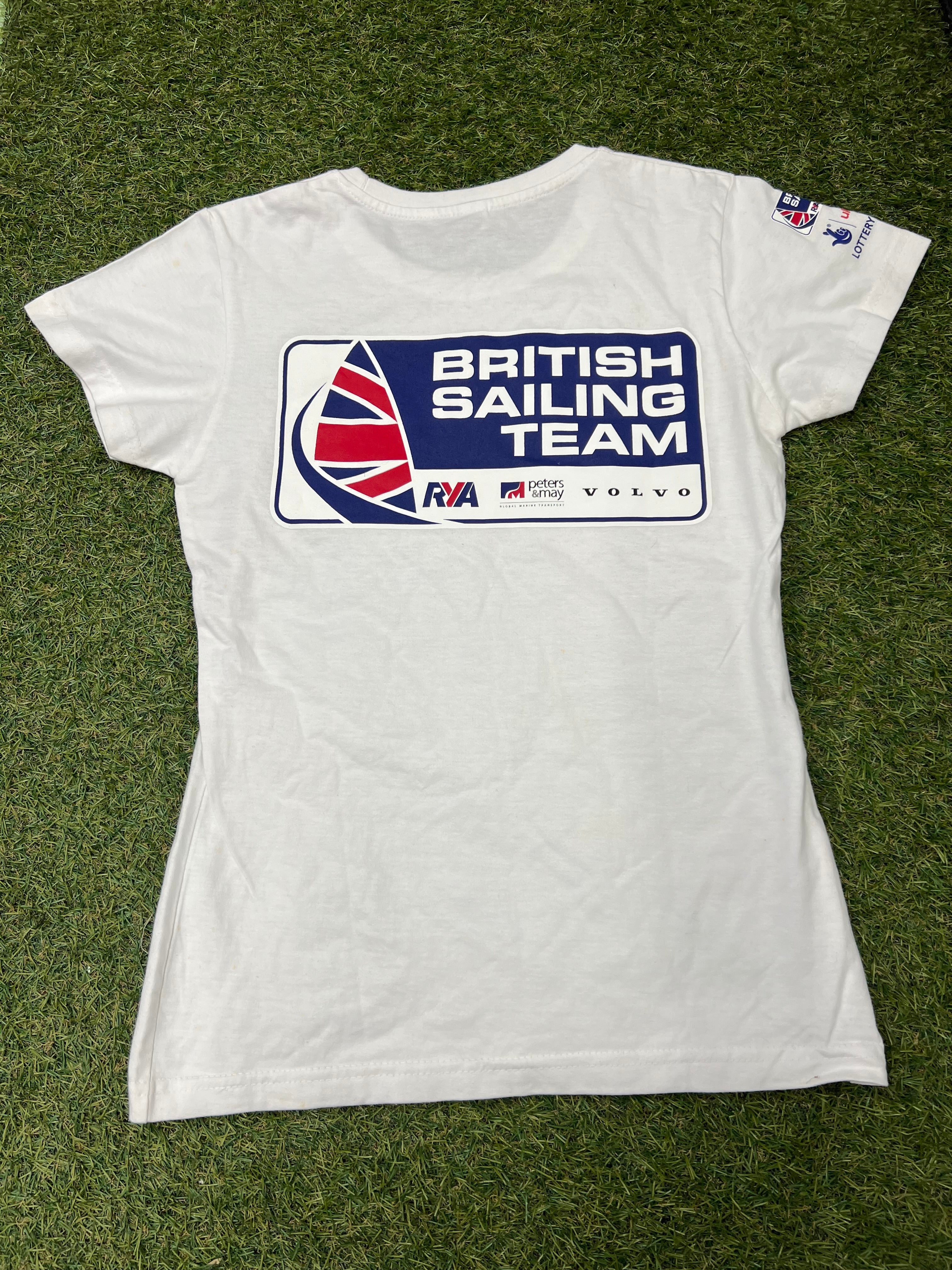 Back view of the white women's British Sailing Team t-shirt showing large blue, red, and white team logo with sponsors on grass background
