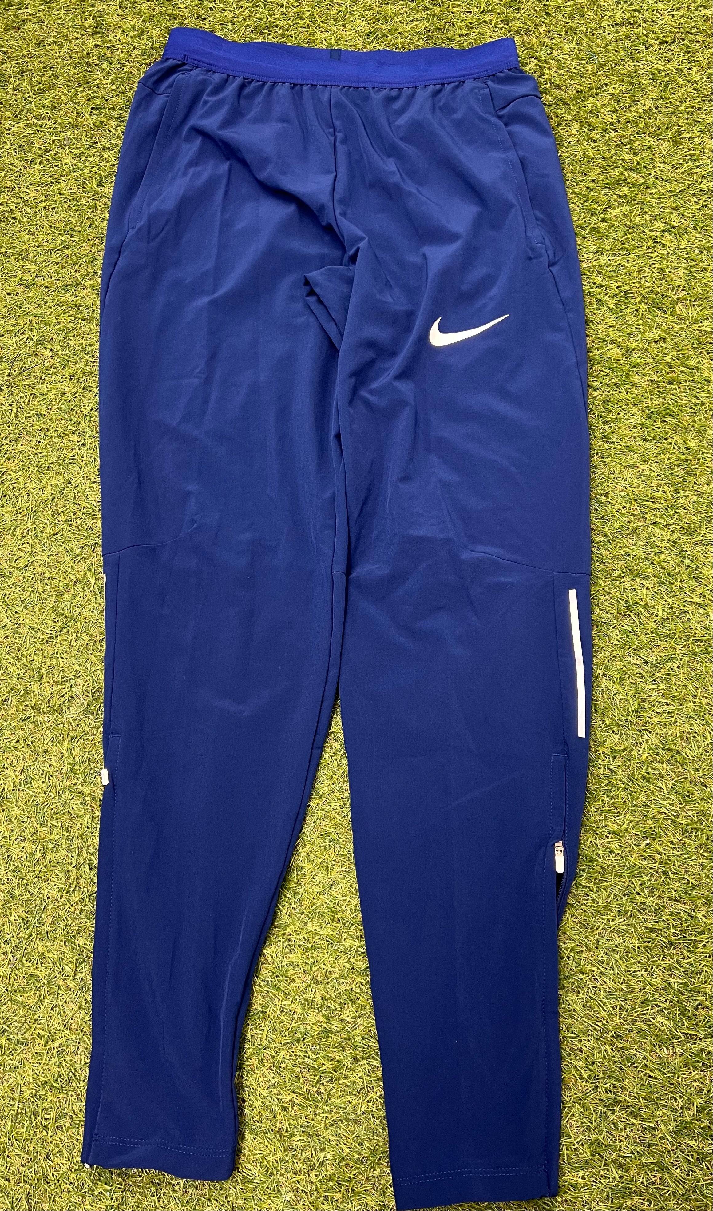 Front view of Nike Women's navy blue Dri-FIT athletic track pants with elastic waistband and white logo on green grass background