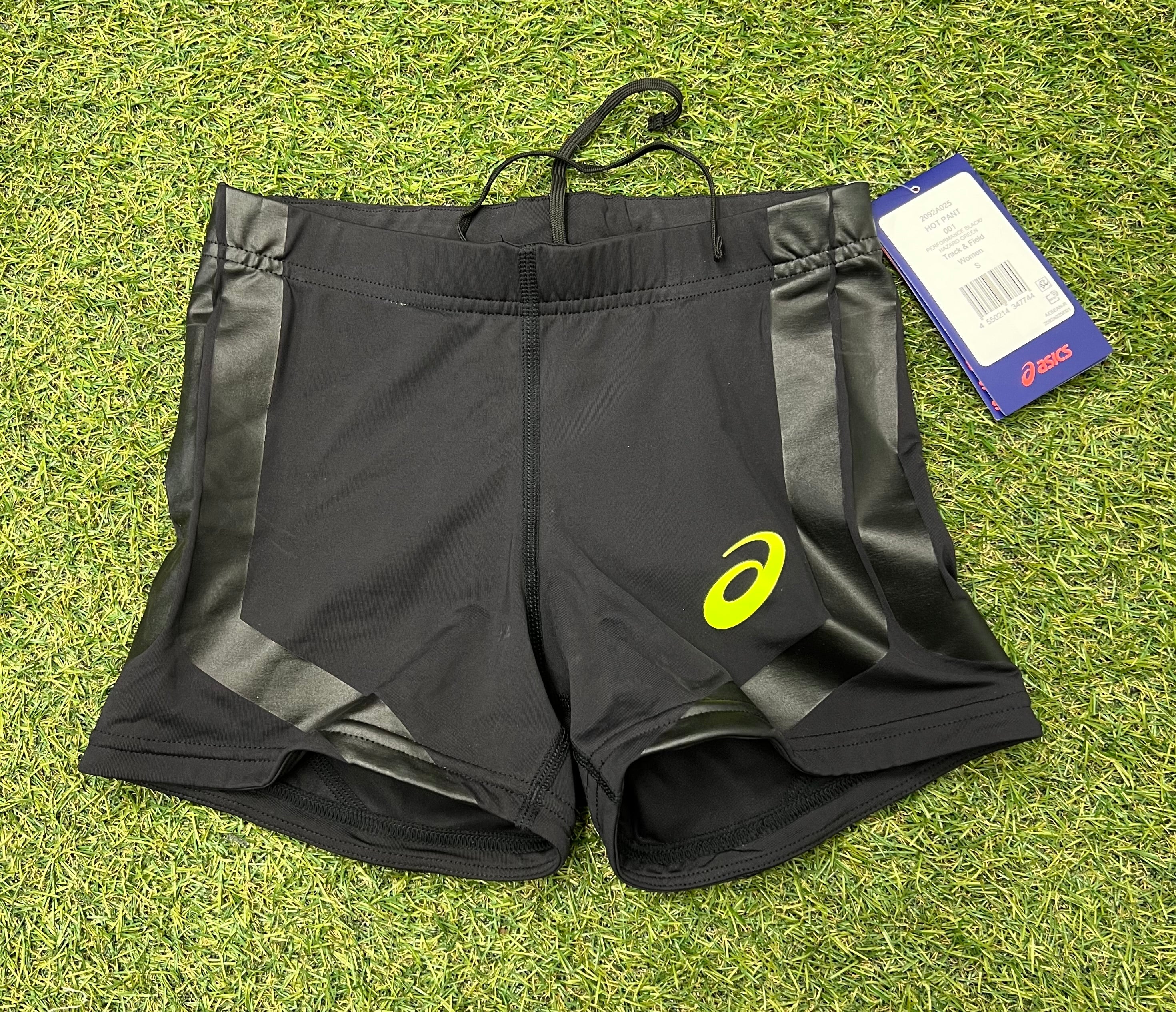Front view of black ASICS women's performance hot pants with hazard green logo and side panels on artificial grass