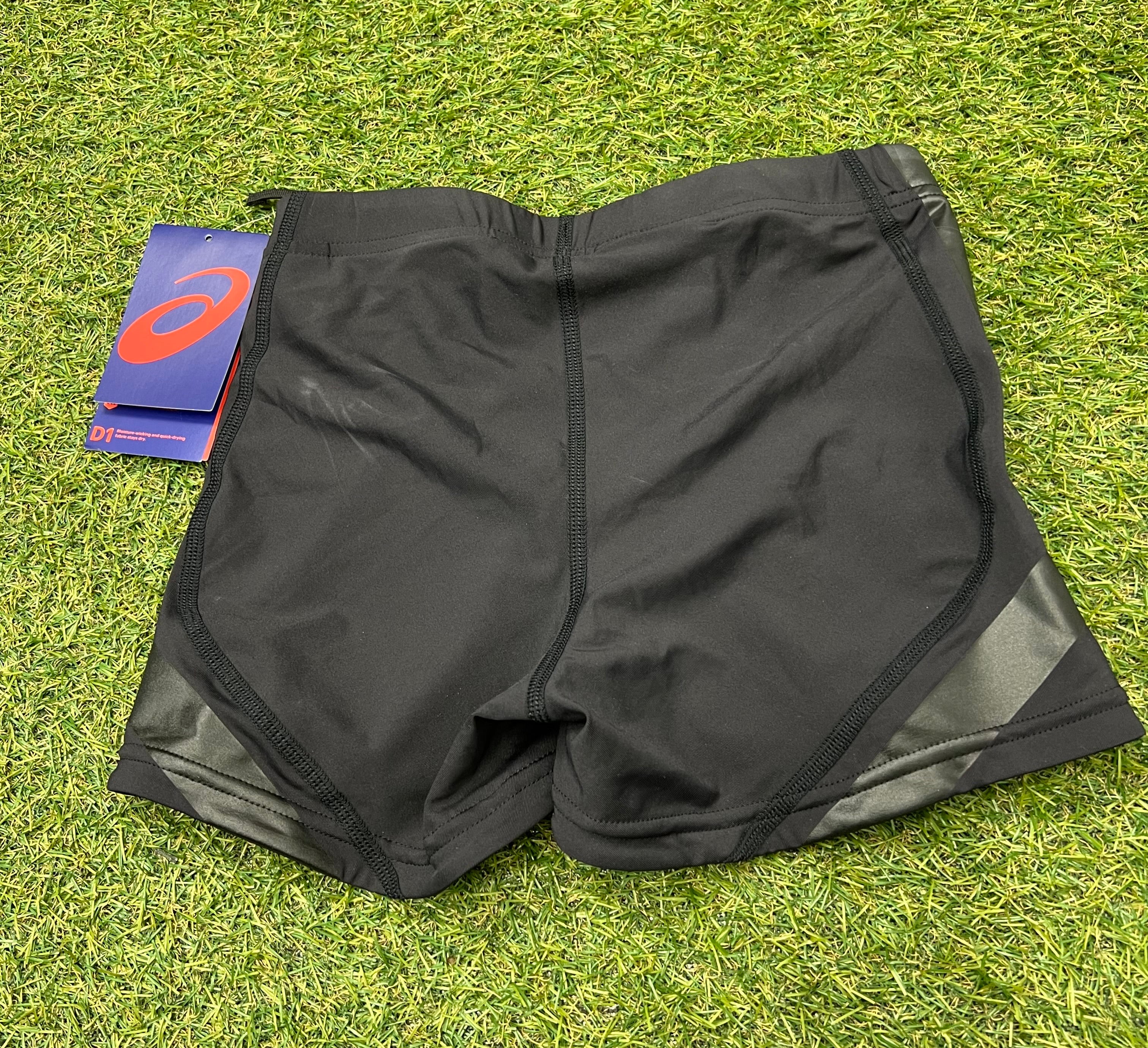 Back view of black ASICS women's performance hot pants showing seamless stitching and side glossy panels on artificial grass