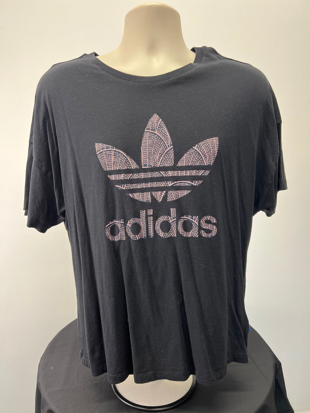 Front view of black Adidas t-shirt featuring pink patterned trefoil logo and brand name printed across chest