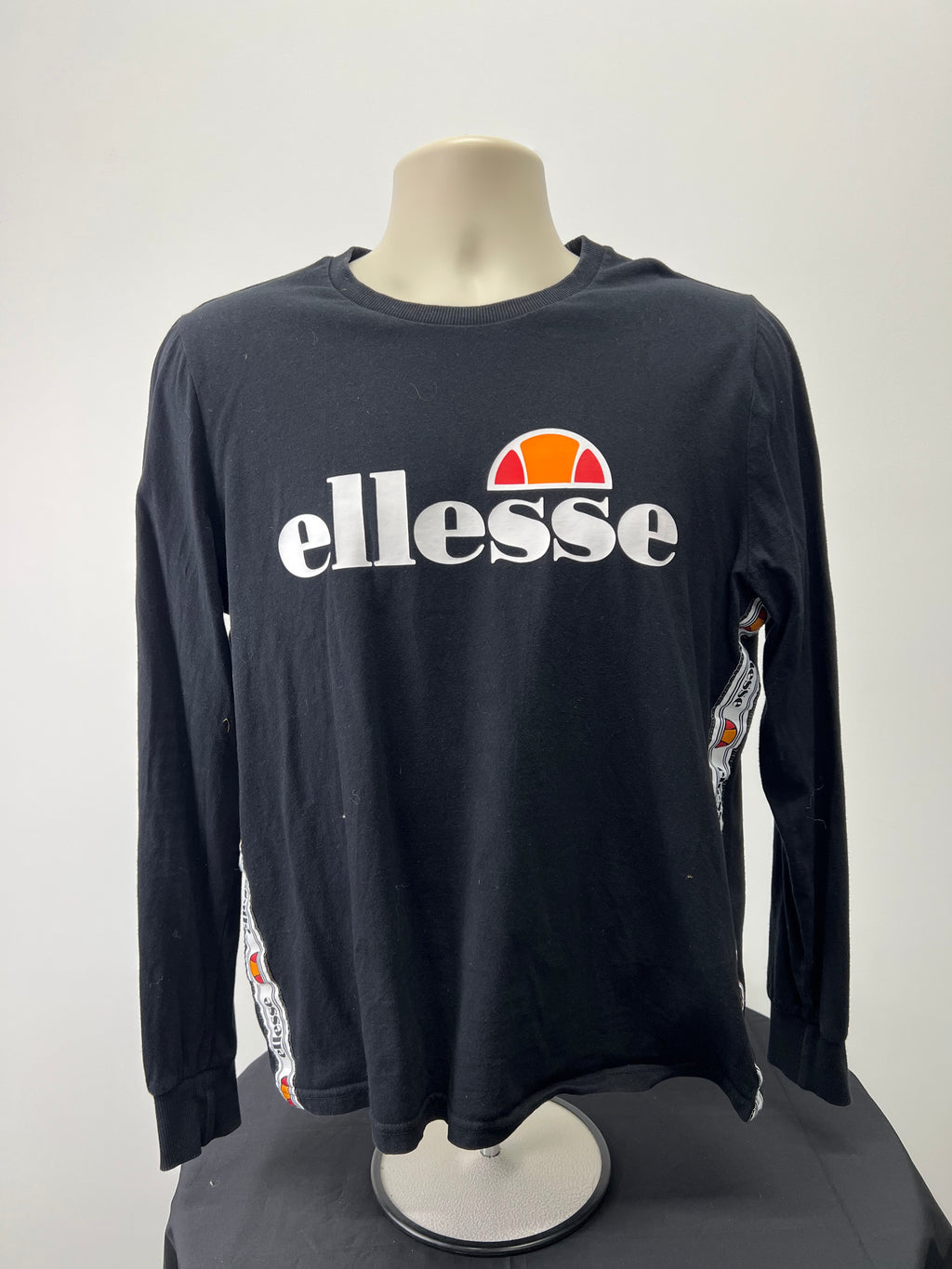 Front view of black ellesse women's long sleeve top with large white ellesse logo and orange-red half-ball graphic on the chest, side logo taping detail