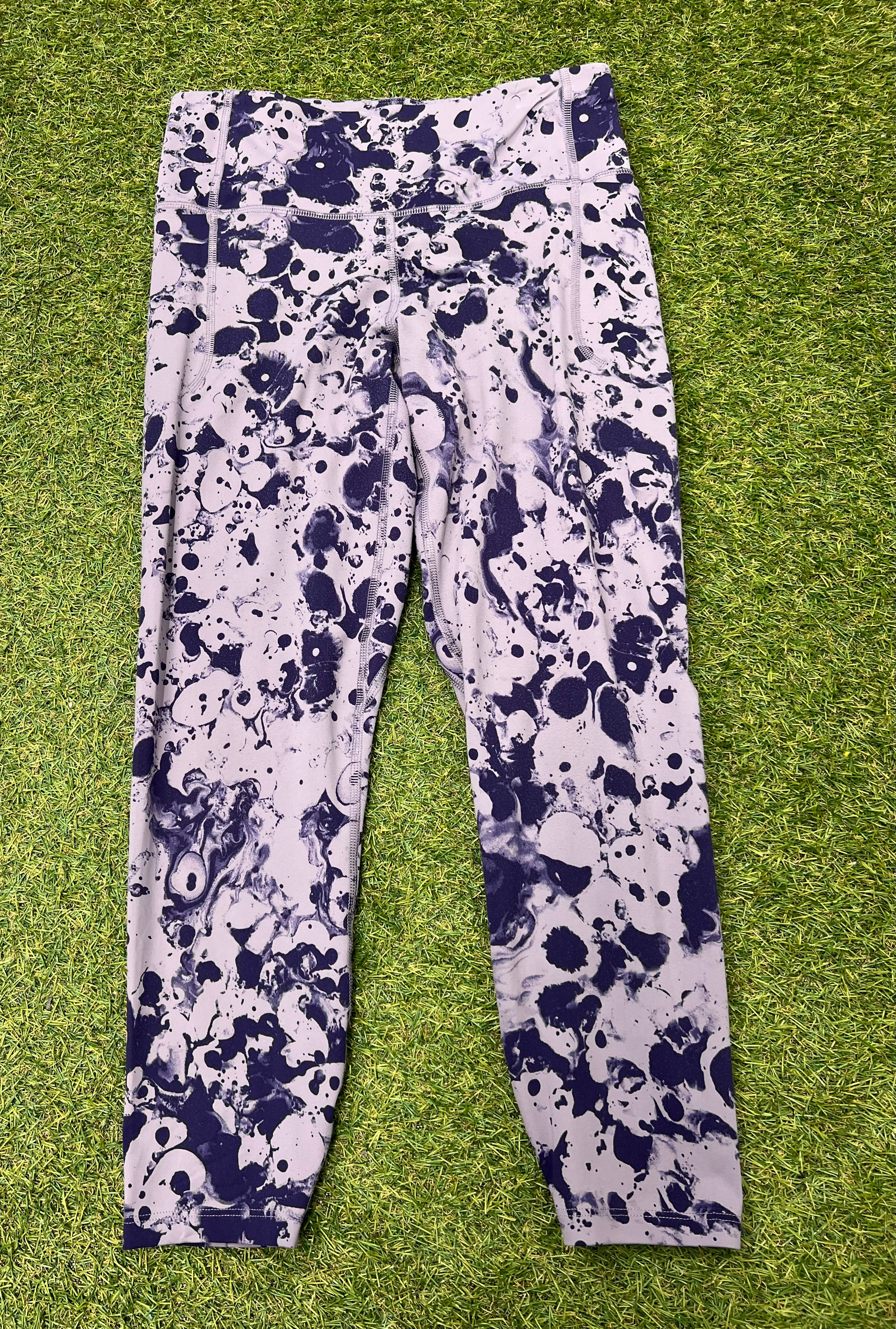 Front view of Under Armour fitted leggings with a black and white splash pattern on lavender background laid on grass