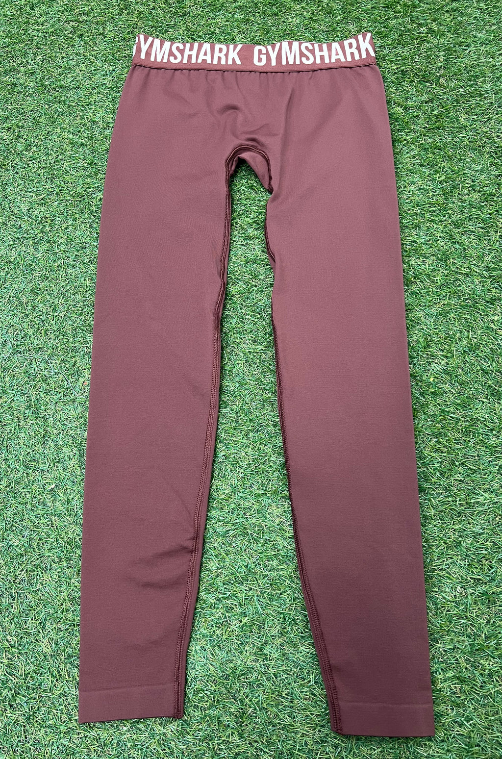 Front view of maroon Gymshark leggings laid flat on grass showing elastic waistband with Gymshark logo