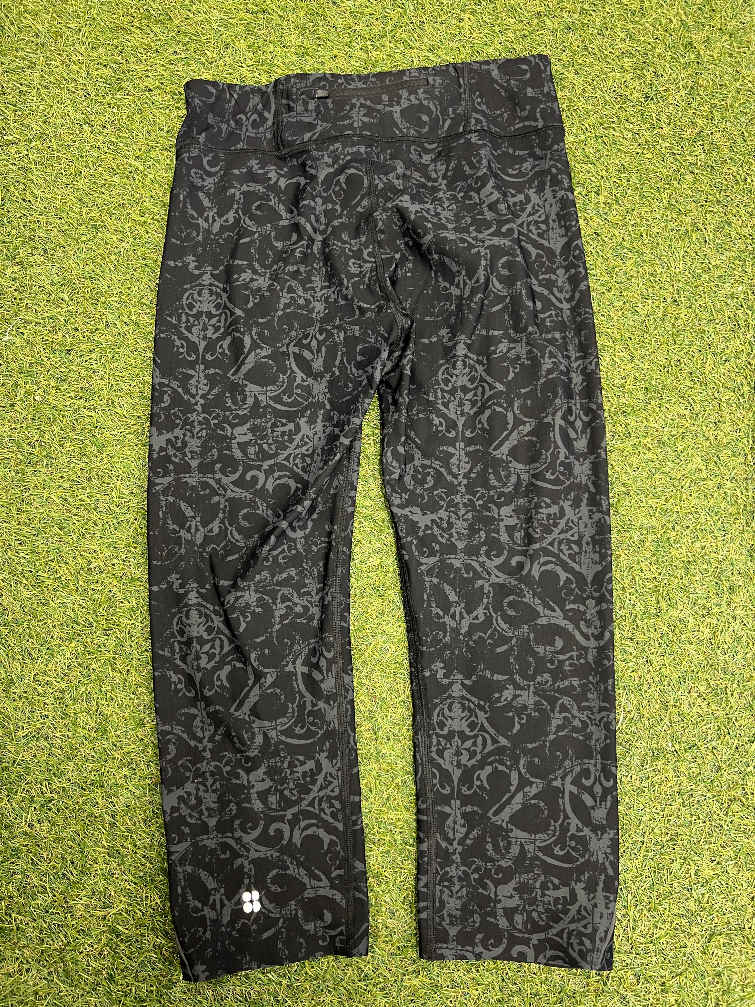 Back view of black and grey printed Sweaty Betty 7/8 leggings showing reflective detail on lower leg, lying on grass