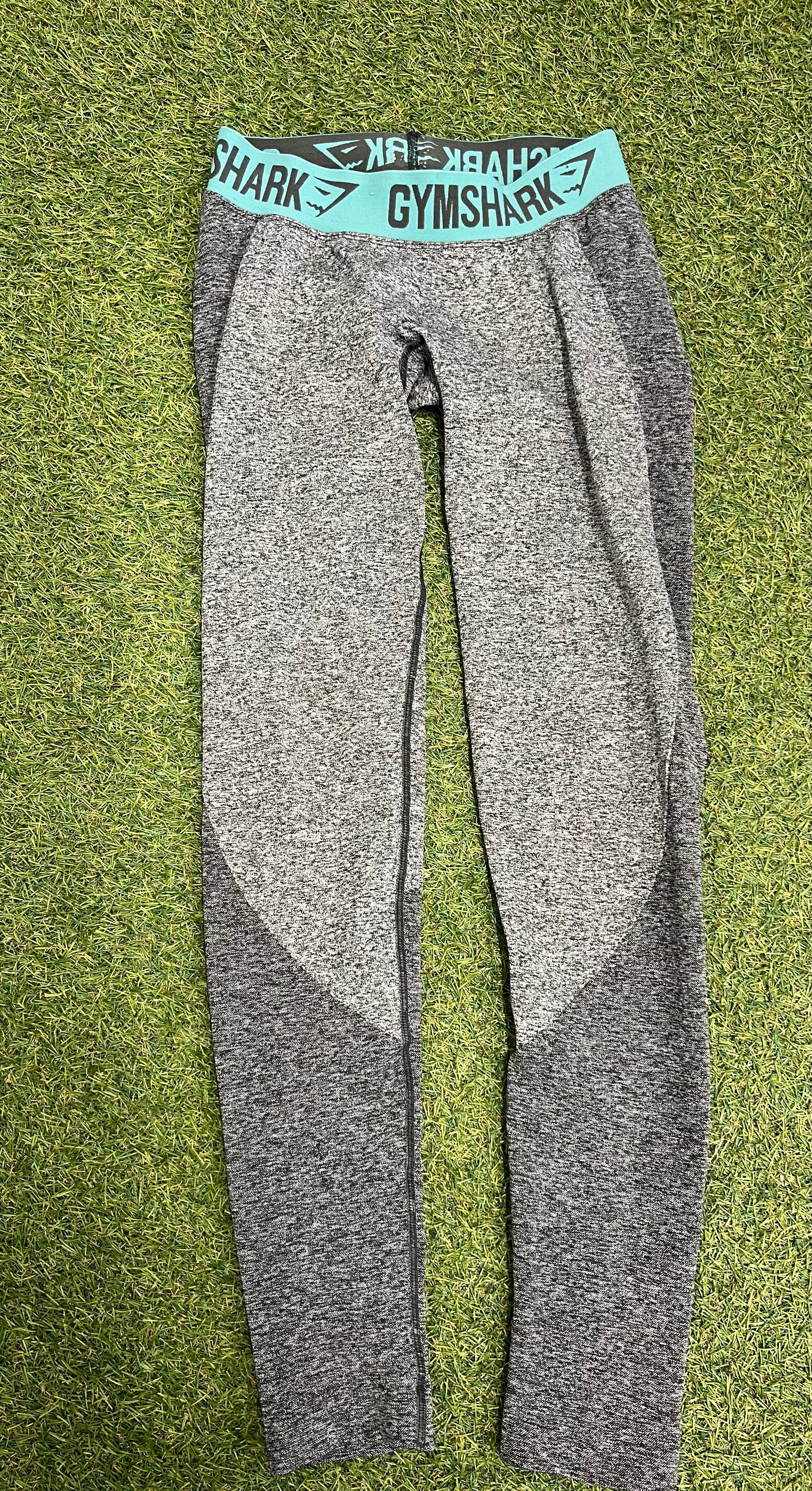 Front view of grey Gymshark seamless leggings with turquoise branded elastic waistband laid flat on green grass