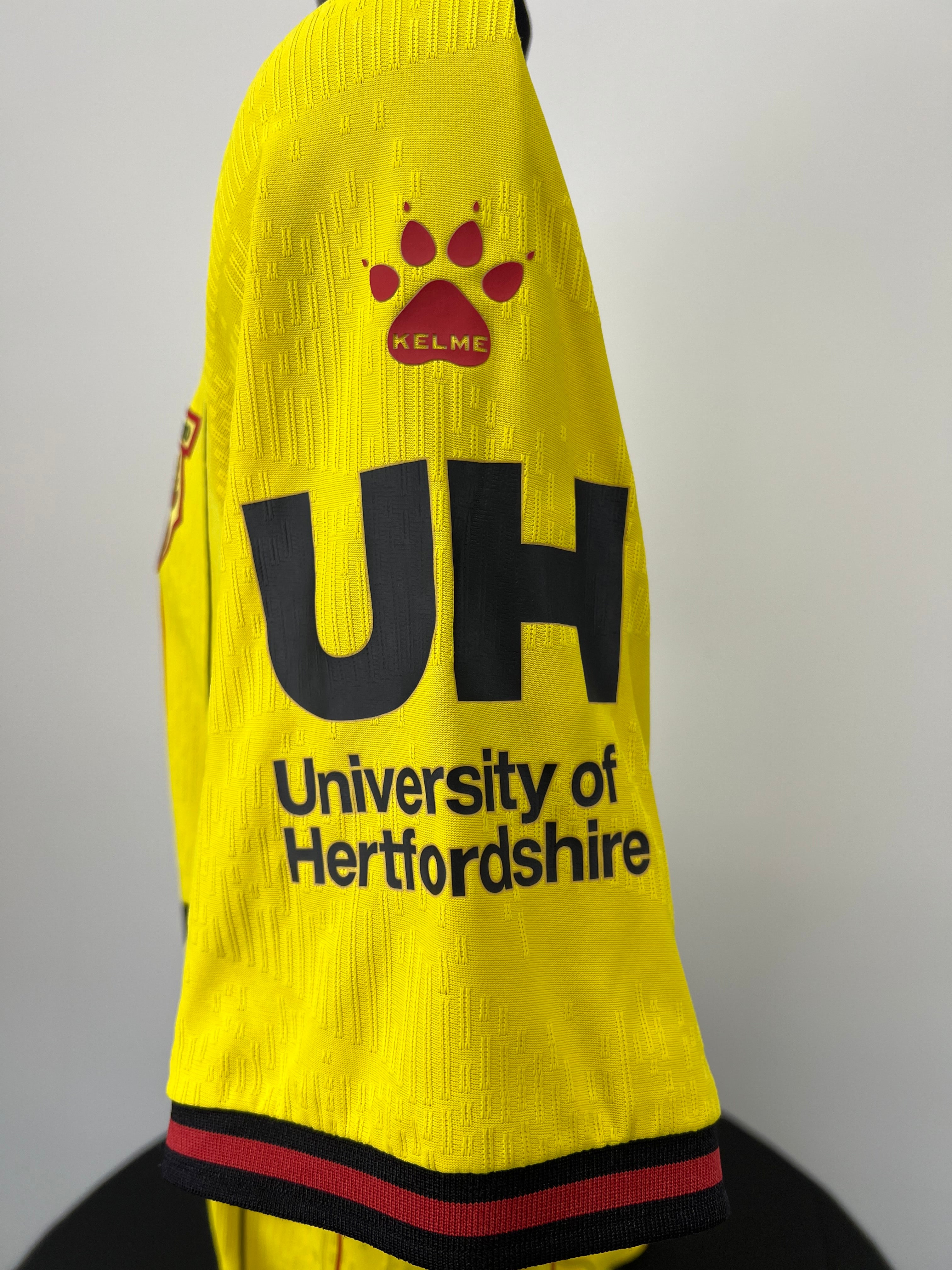Close-up of right sleeve featuring Kelme paw print logo and University of Hertfordshire sponsor in black on yellow fabric