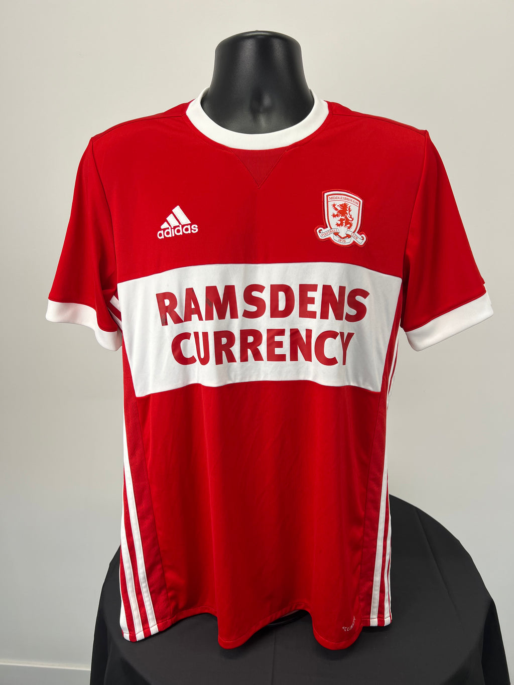 Front view of a Middlesbrough FC red football shirt with white horizontal band, Adidas logo, and Ramsdens Currency sponsor logo