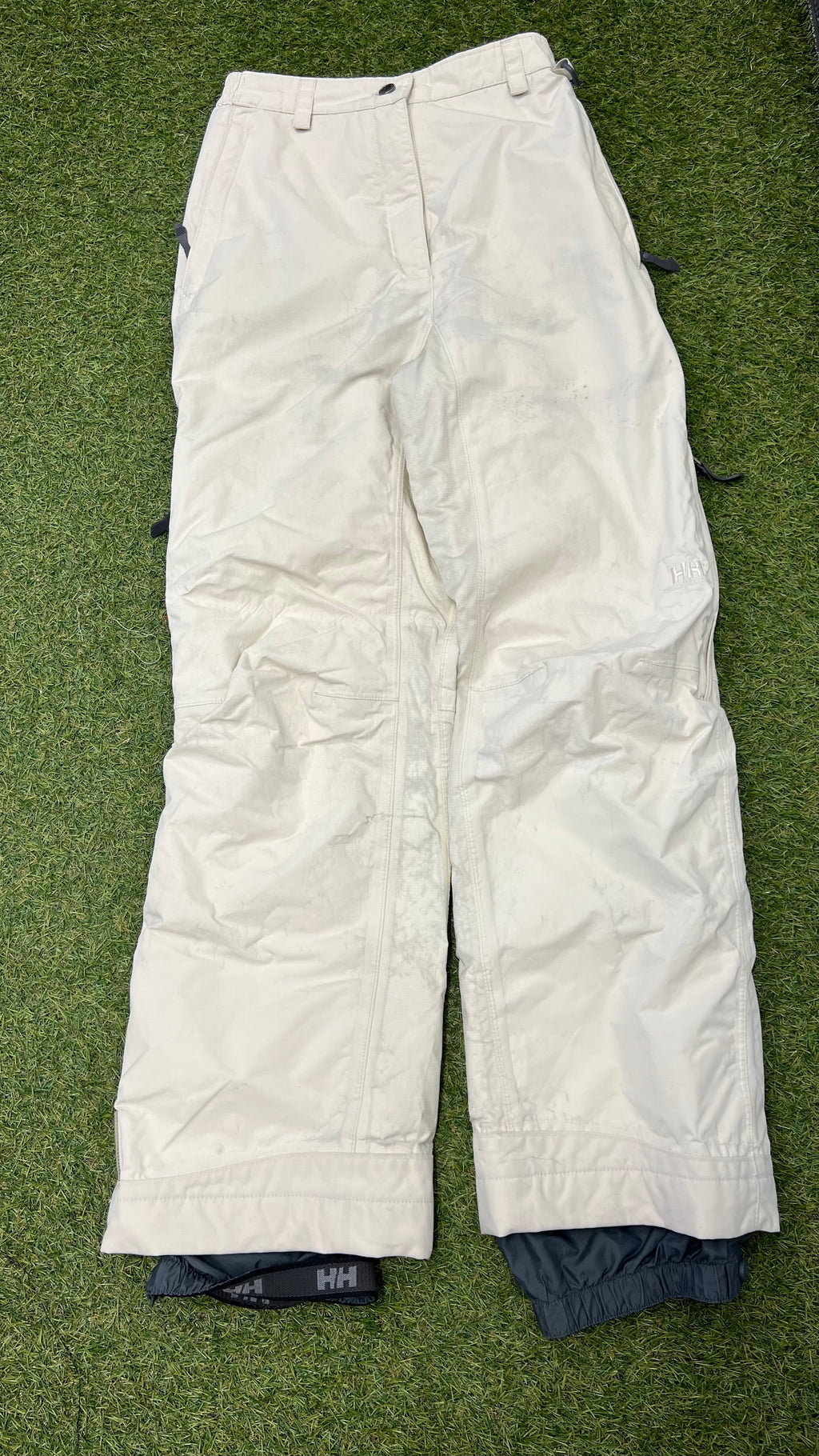Front view of cream Helly Hansen women's ski trousers laid flat on grass showing button and zip closure with side adjustment straps