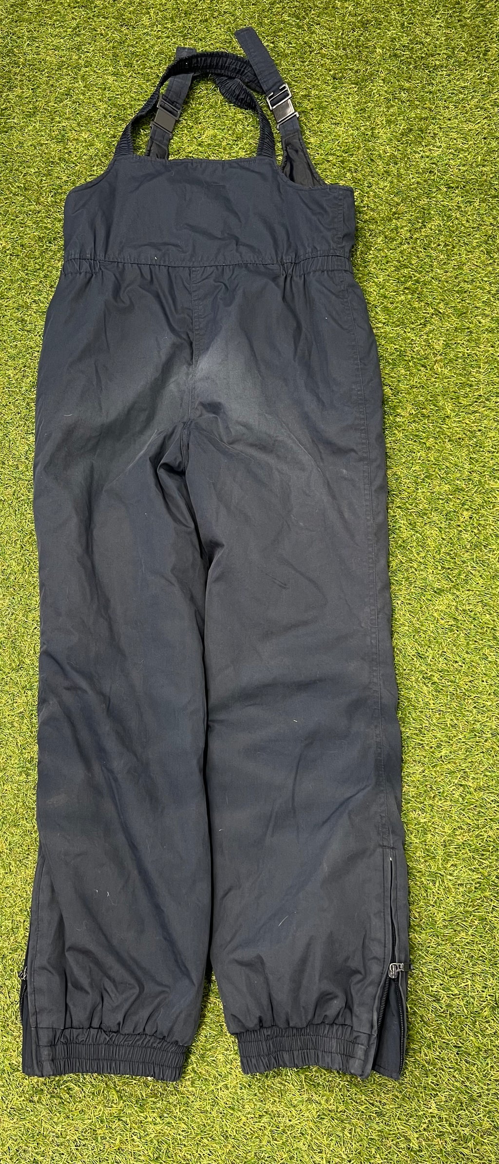 Back view of black Campri Ski Action insulated salopettes showing adjustable shoulder straps and ankle zippers laid on grass background