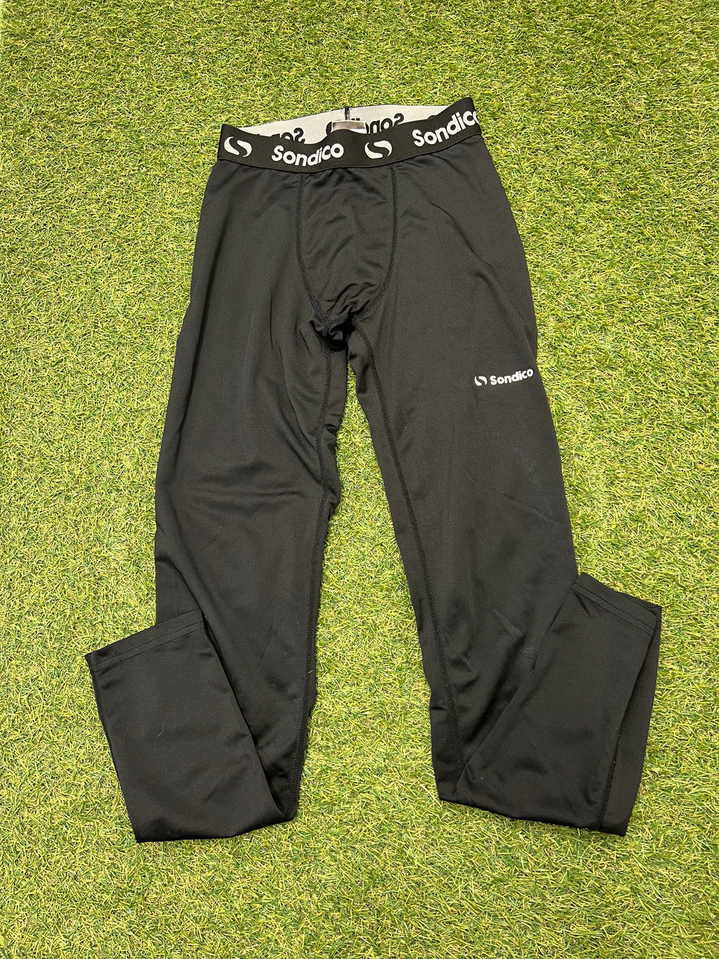 Close-up view of black Sondico kids leggings with elastic waistband and logo on grassy surface.