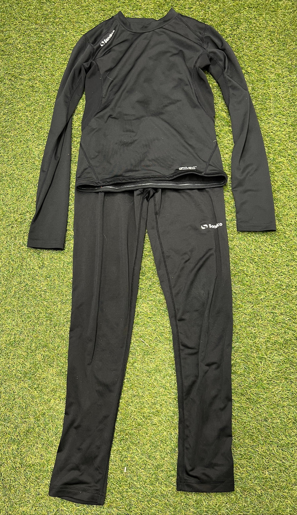 Full view of black Sondico kids base layer set with long sleeve top and leggings laid flat on grass