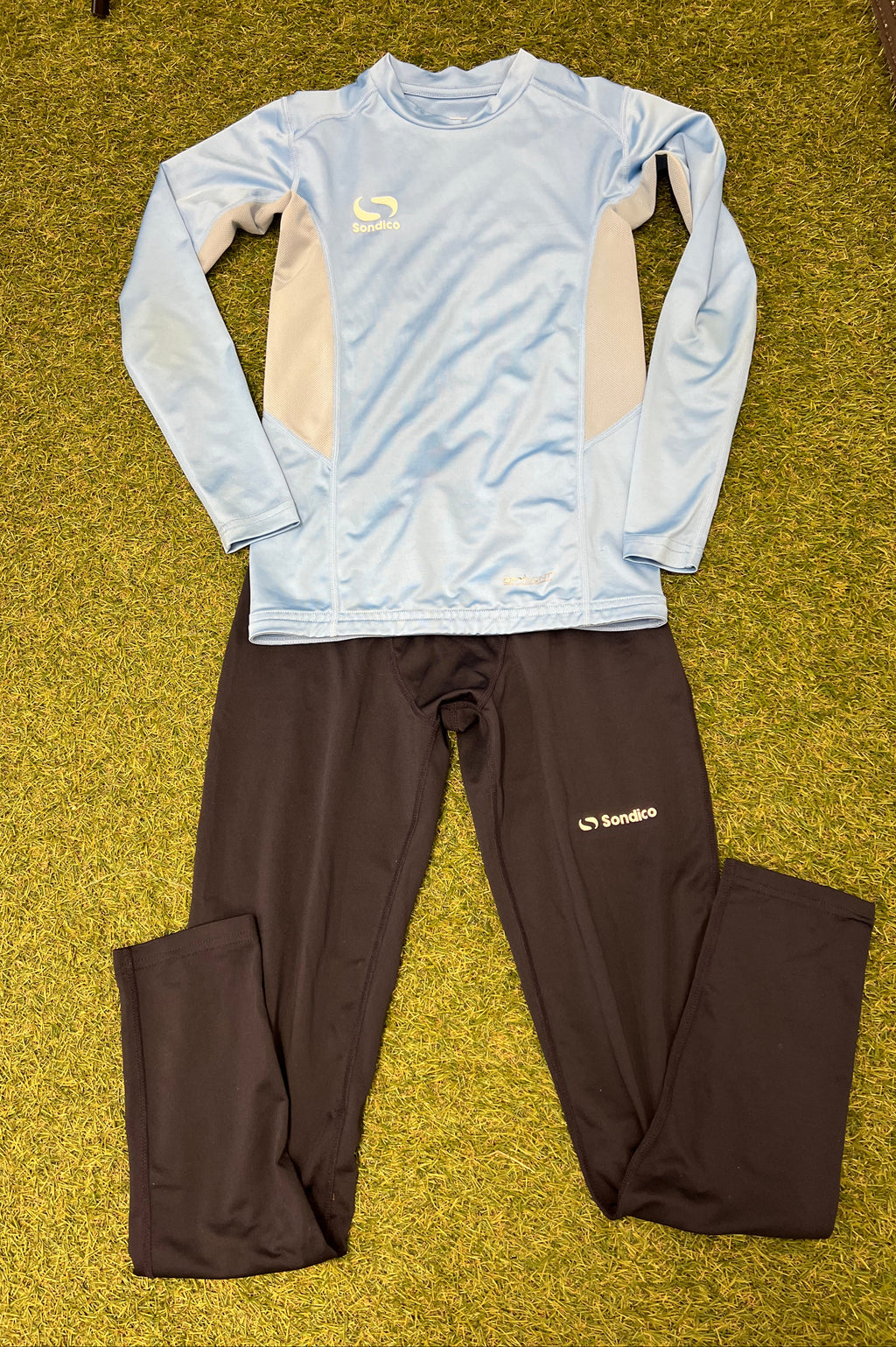 Sondico kids base layer set laid out on grass showing a sky blue long-sleeve top with beige mesh side panels and black leggings
