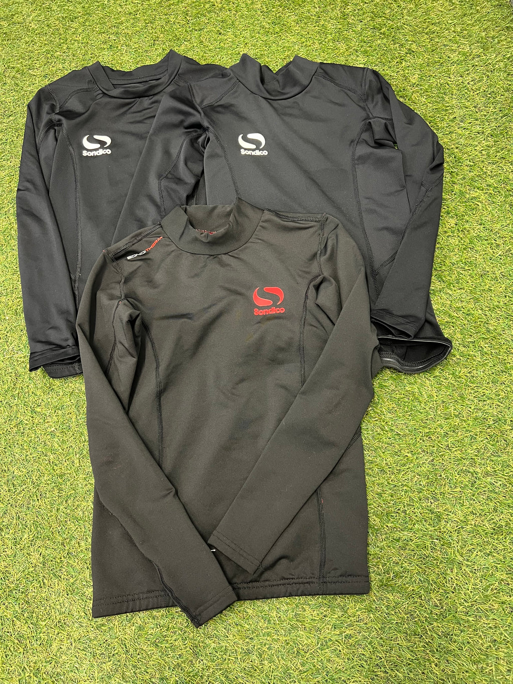 Three Sondico kids' thermal base layer tops in black laid flat on grass, two with white logos, one with a red logo