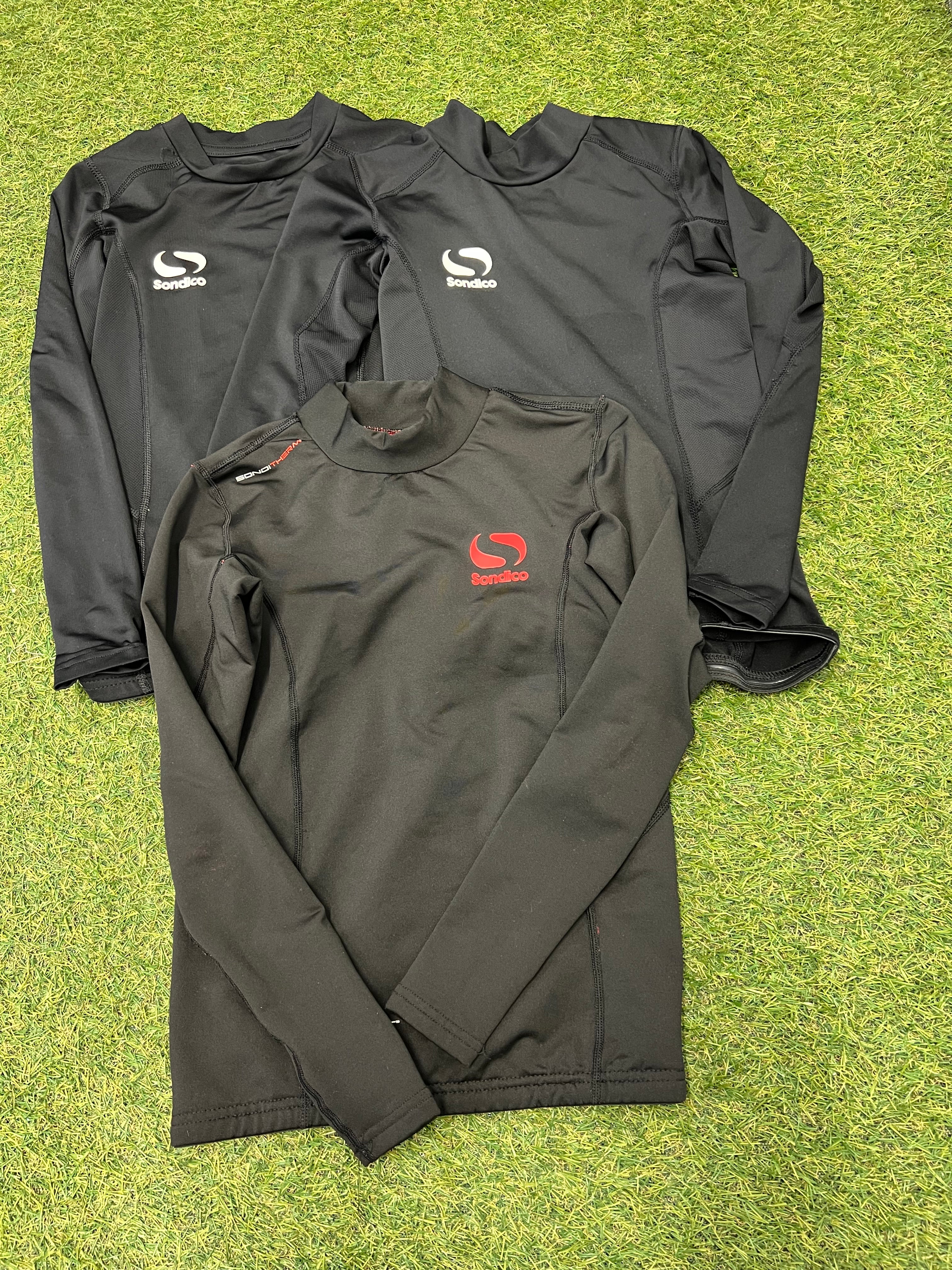 Three Sondico kids' thermal base layer tops in black laid flat on grass, two with white logos, one with a red logo