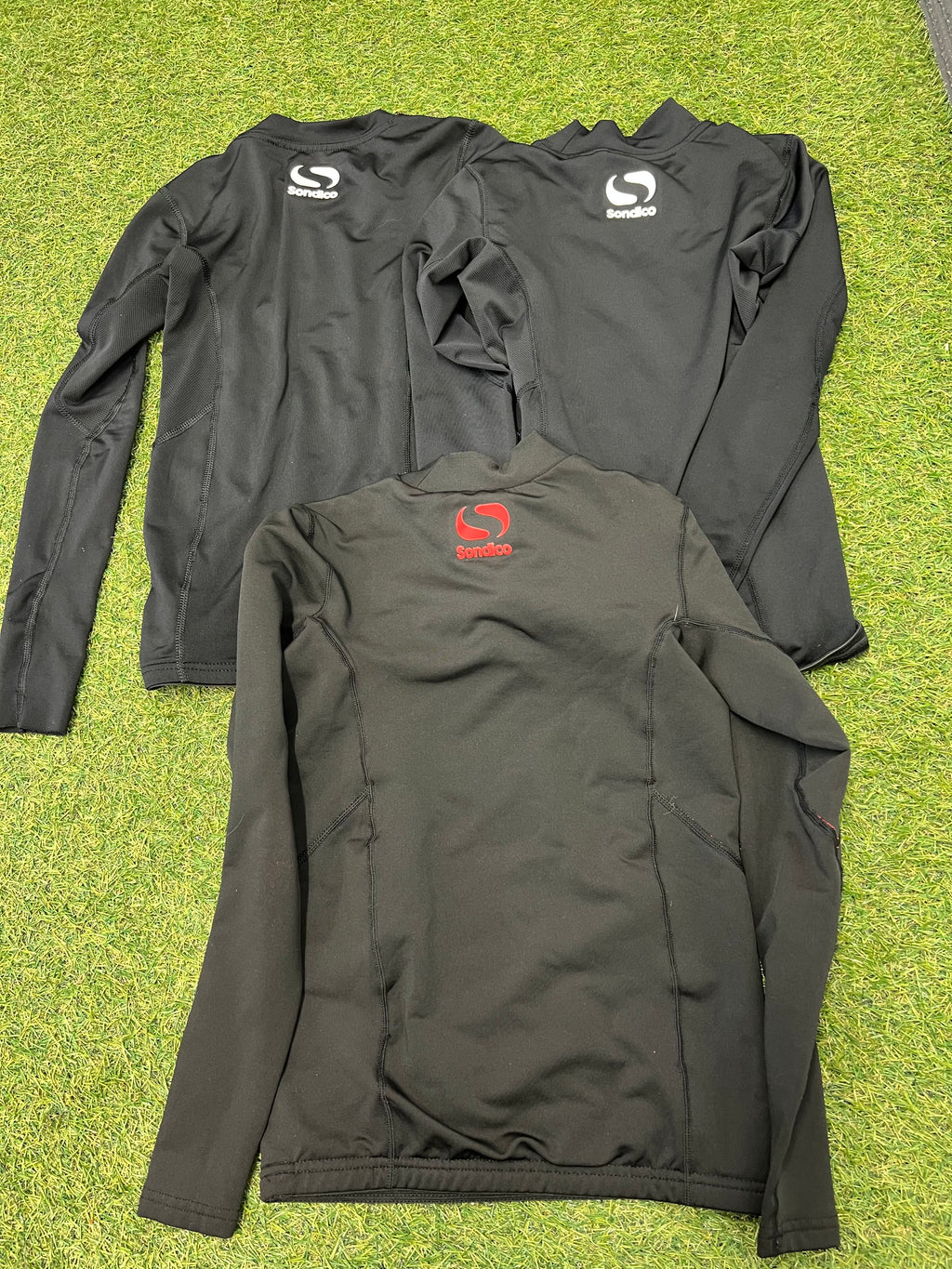 Back view of three Sondico kids' black thermal base layer tops laid flat on grass, showing white and red logos on upper backs