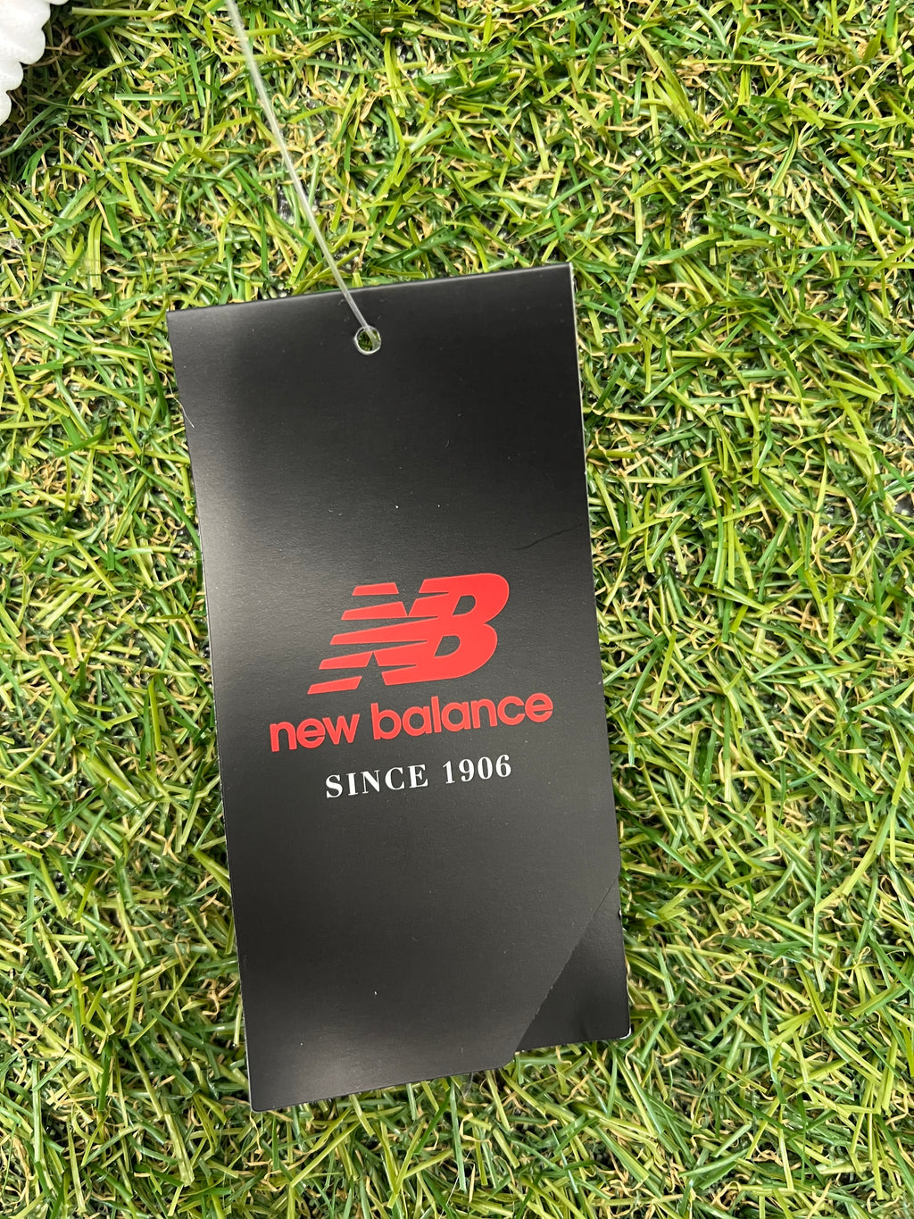 Black New Balance brand tag with red logo and 'Since 1906' text on green grass background