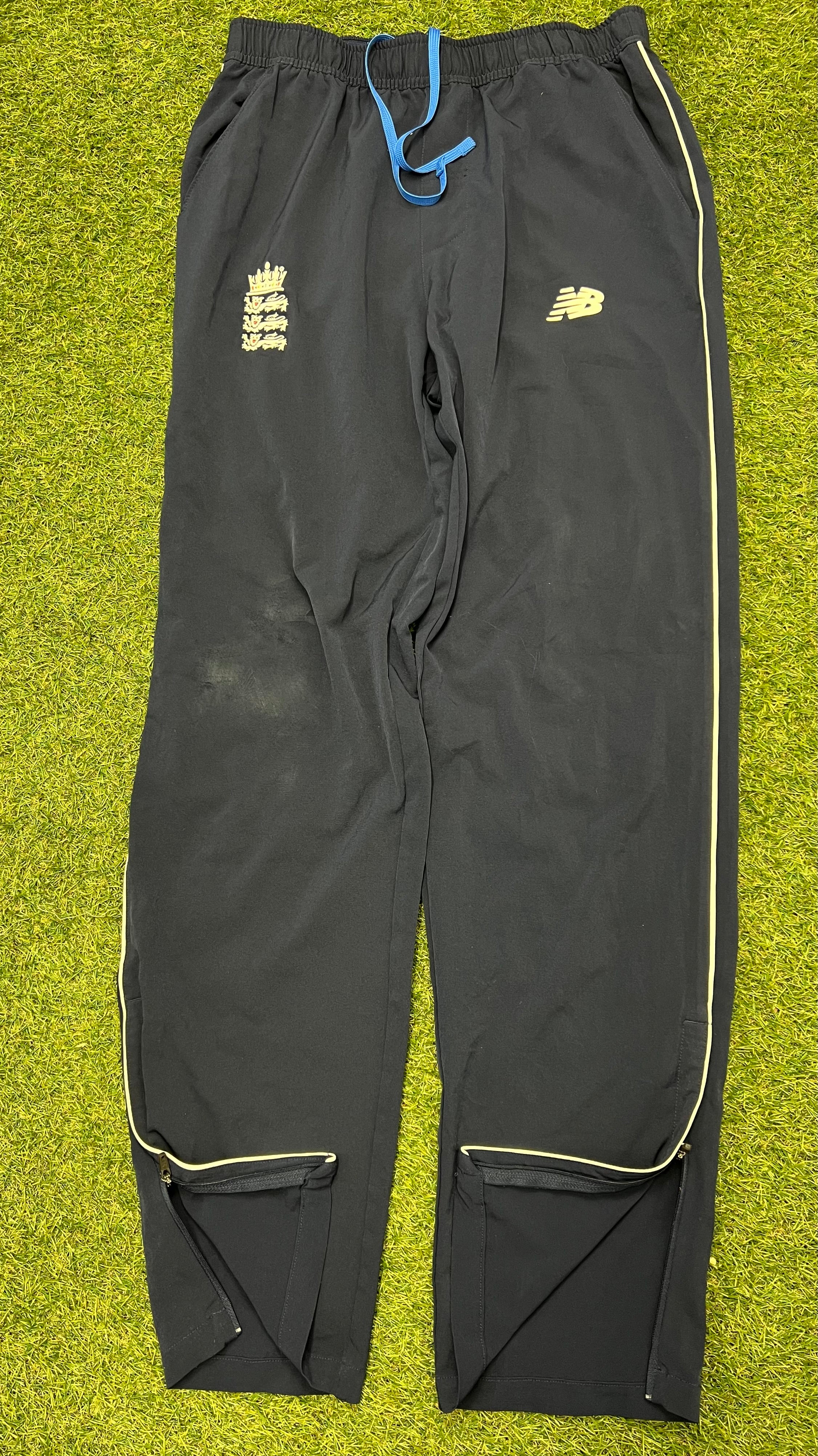 Front view of black men’s New Balance cricket tracksuit bottoms with yellow piping and blue drawstring on green grass