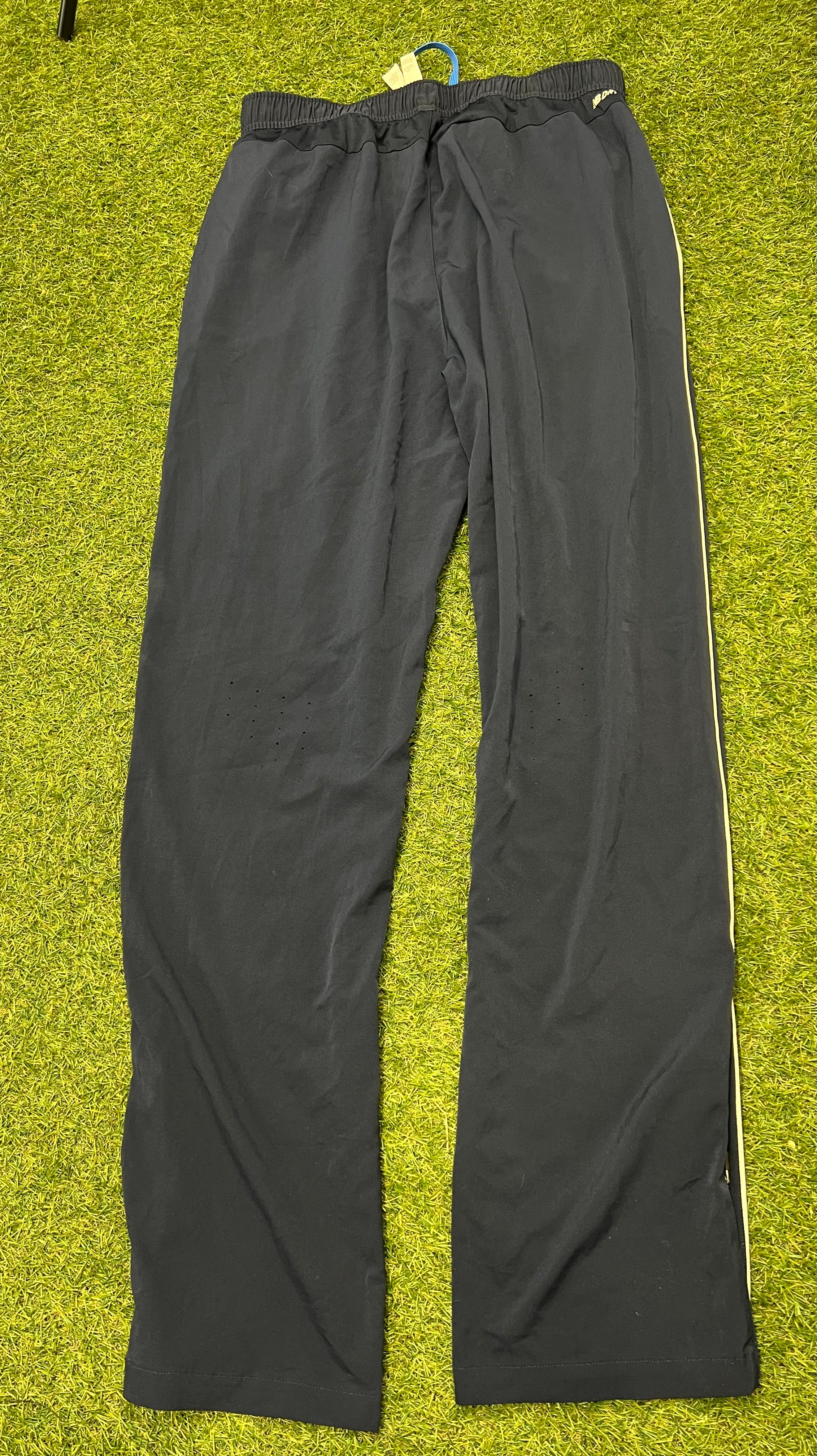 Rear view of black men’s New Balance cricket tracksuit bottoms laid flat on green grass showing plain back and elastic waistband