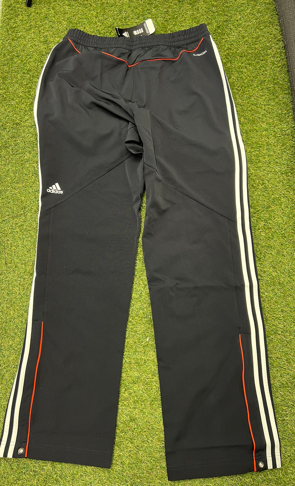 Rear view of black Adidas training trousers showing Adidas logo and red piping detail on grass background