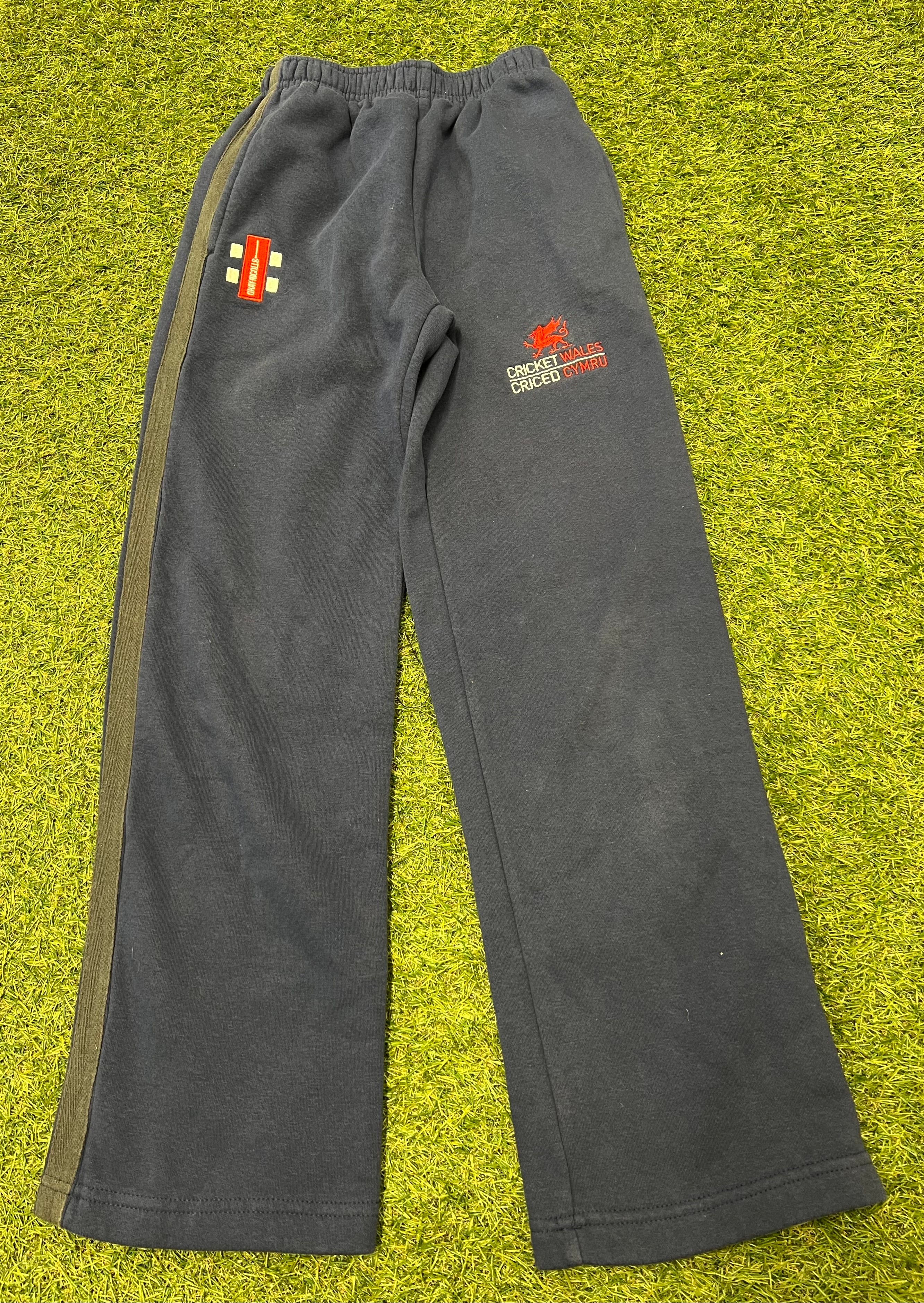 Gray Nicolls kids cricket tracksuit bottoms with Cricket Wales logo laid flat on grass surface