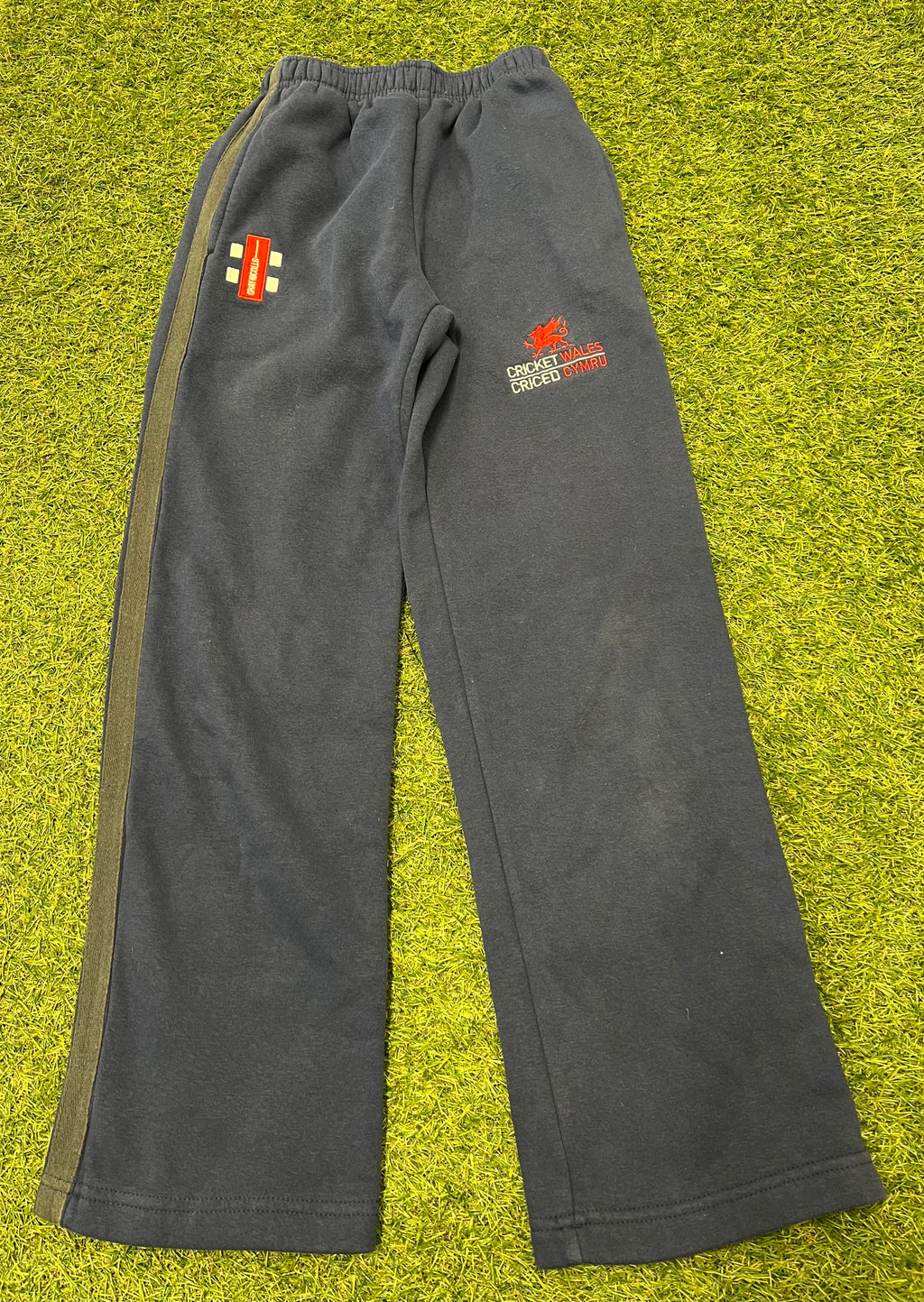 Front view of kids gray Nicholls Cricket Wales tracksuit bottoms with red logo and olive side stripe on grass background