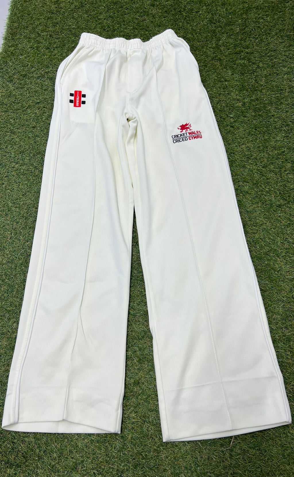 Front view of white cricket trousers with Cricket Wales logo and Gray-Nicolls branding on grass background