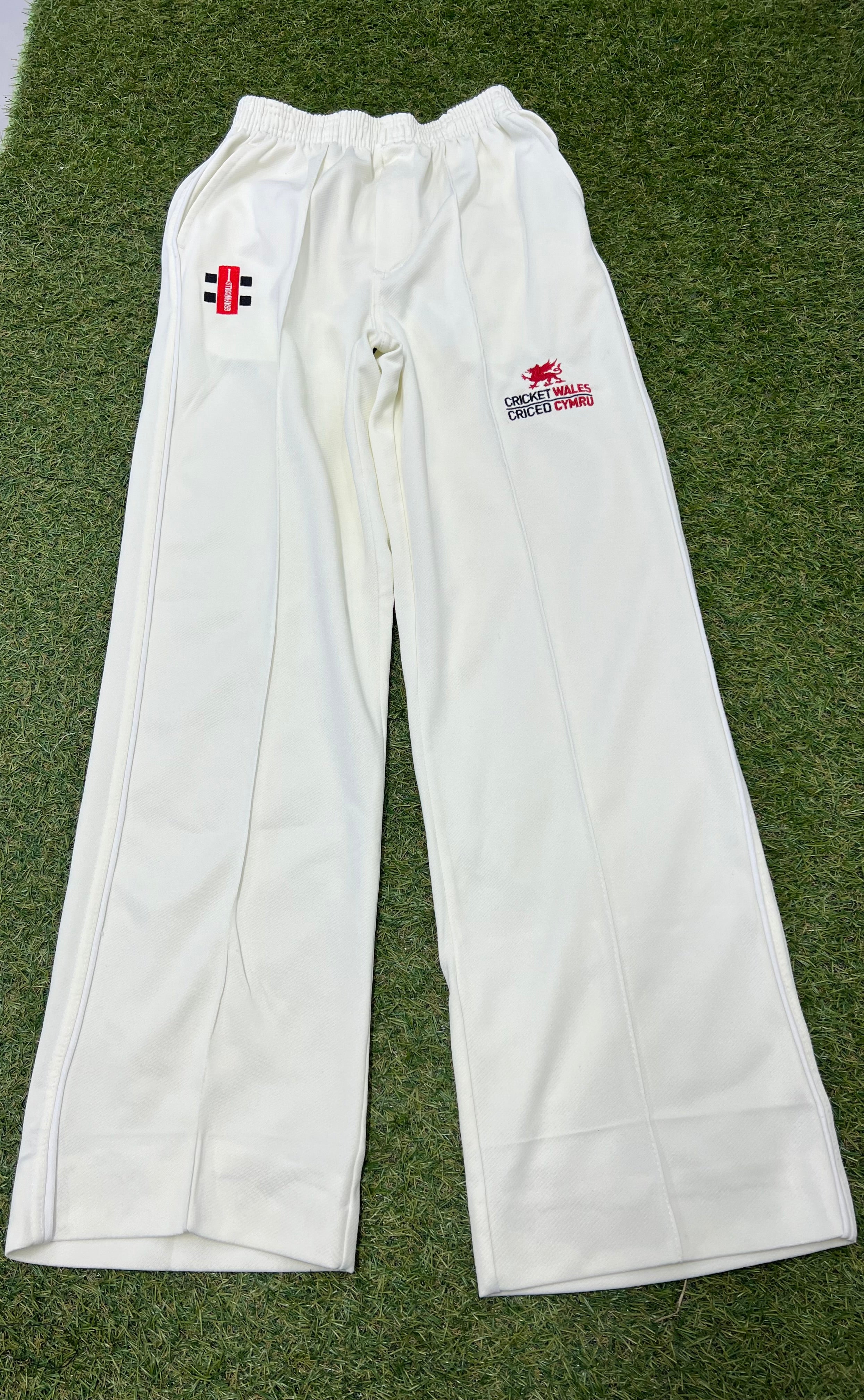 Front view of white cricket trousers with Cricket Wales logo and Gray-Nicolls branding on grass background