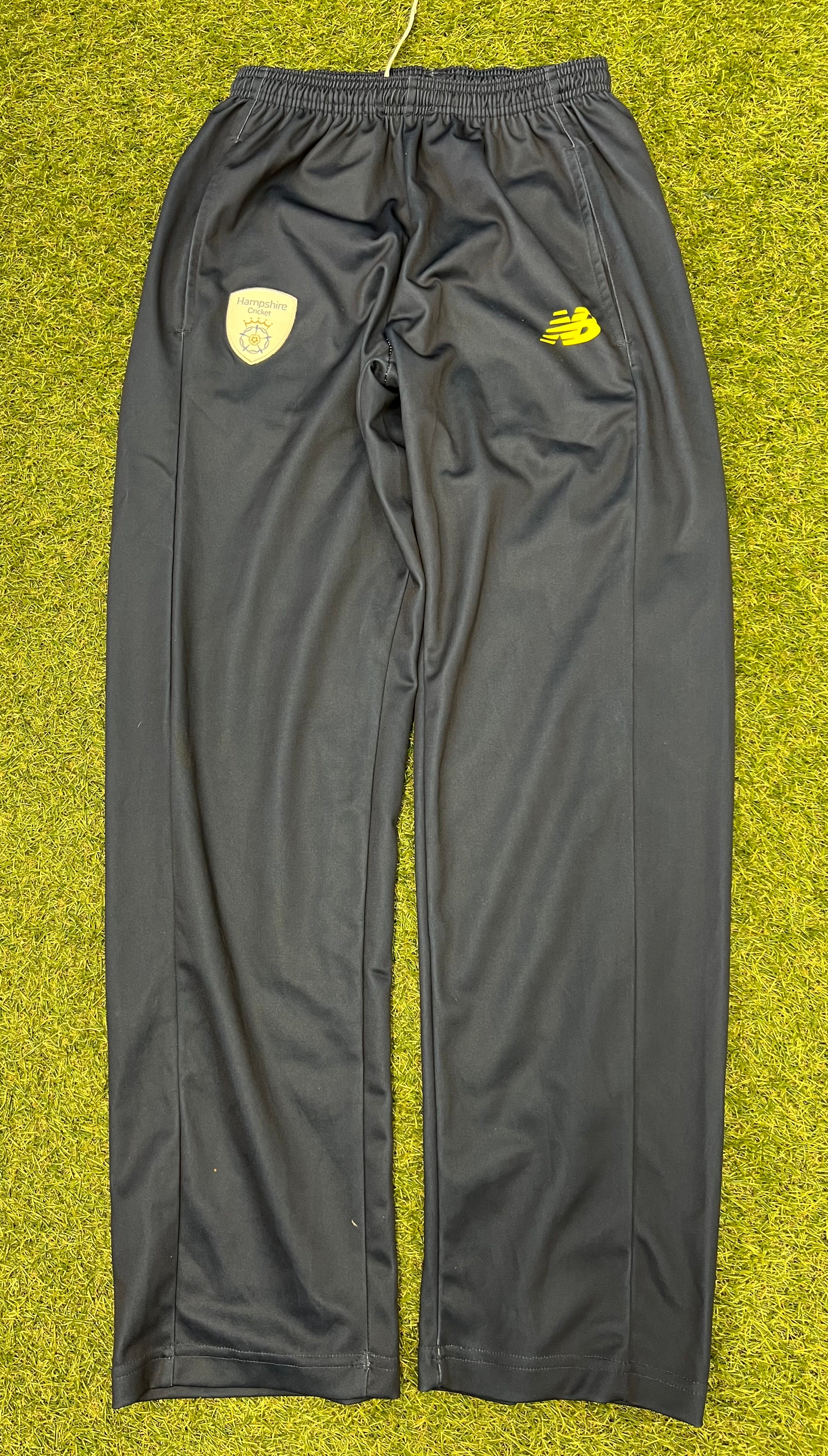 Front view of black New Balance Hampshire cricket tracksuit bottoms with yellow logo and Hampshire Cricket emblem on green turf
