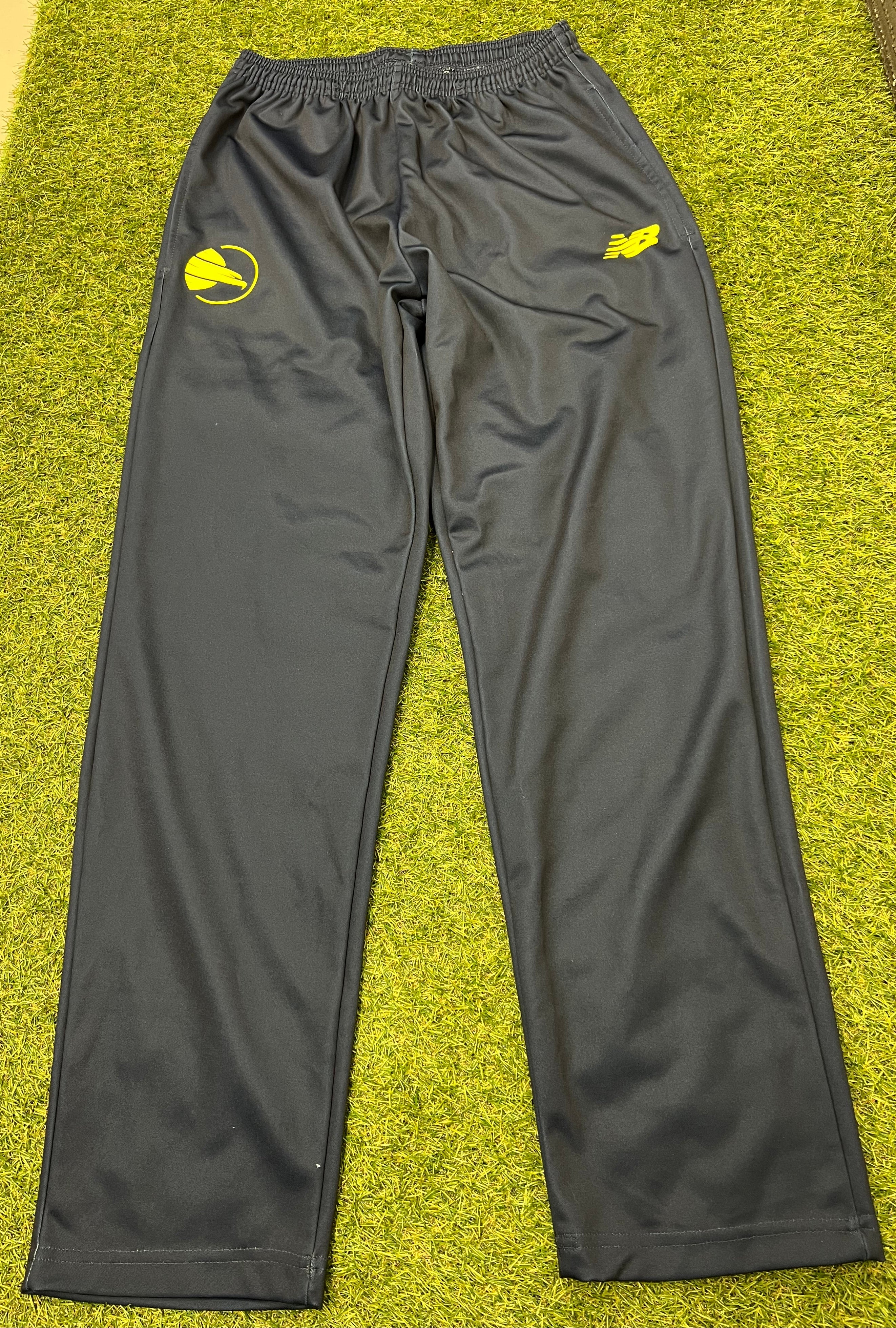 Front view of men's black New Balance tracksuit bottoms with yellow logo on grass background
