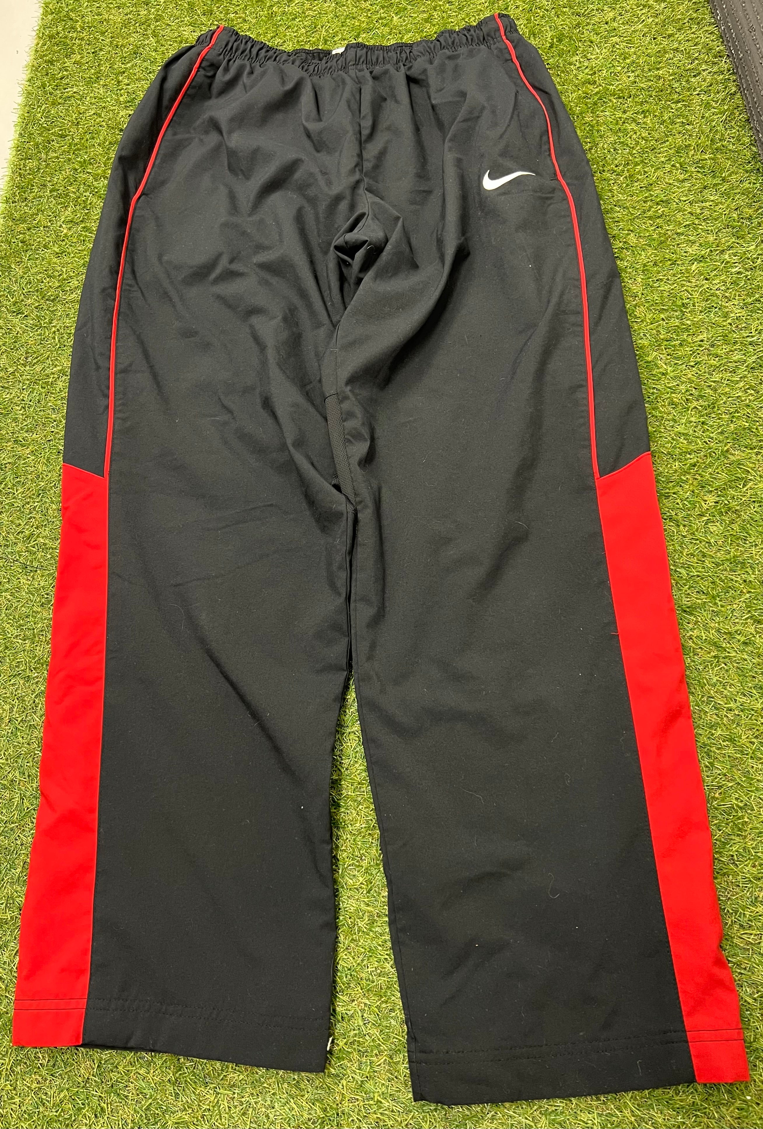 Front view of men's Nike black tracksuit bottoms with red side panels and white Nike logo on left thigh laid flat on grass