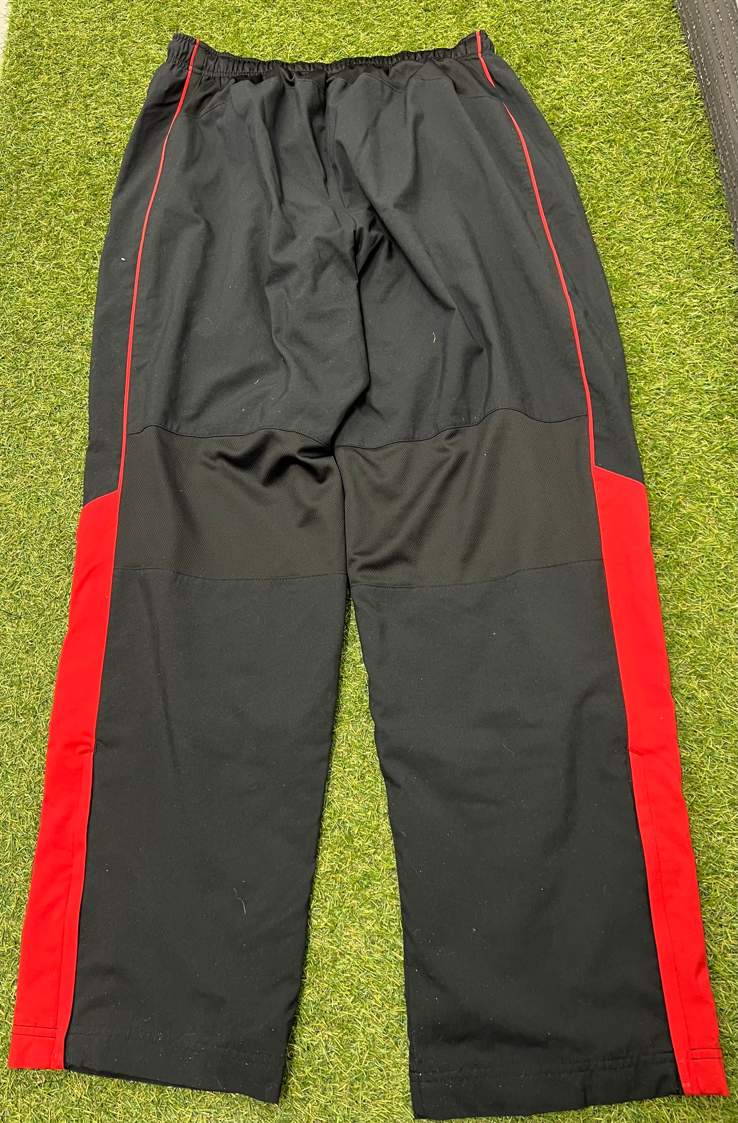 Back view of men's Nike black tracksuit bottoms showing red side panels and black mesh knee area laid flat on grass