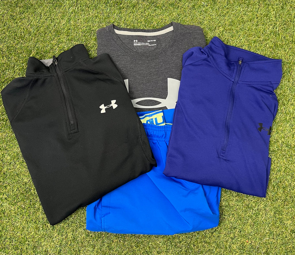 Folded Under Armour men's activewear bundle including black and blue zip-up tops, grey HeatGear t-shirt, and bright blue shorts on grass