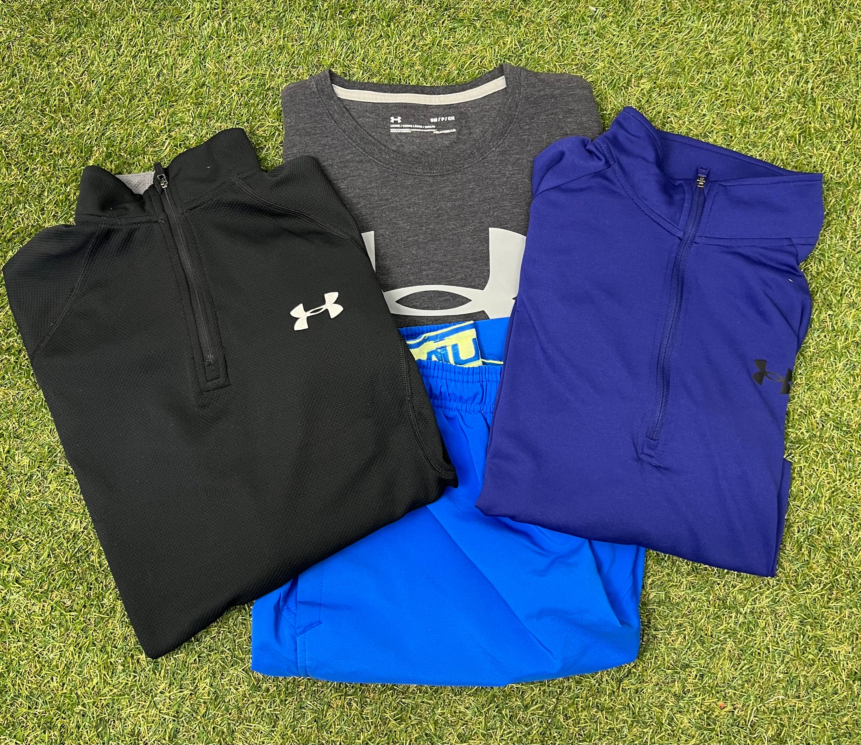 Folded Under Armour men's activewear bundle including black and blue zip-up tops, grey HeatGear t-shirt, and bright blue shorts on grass