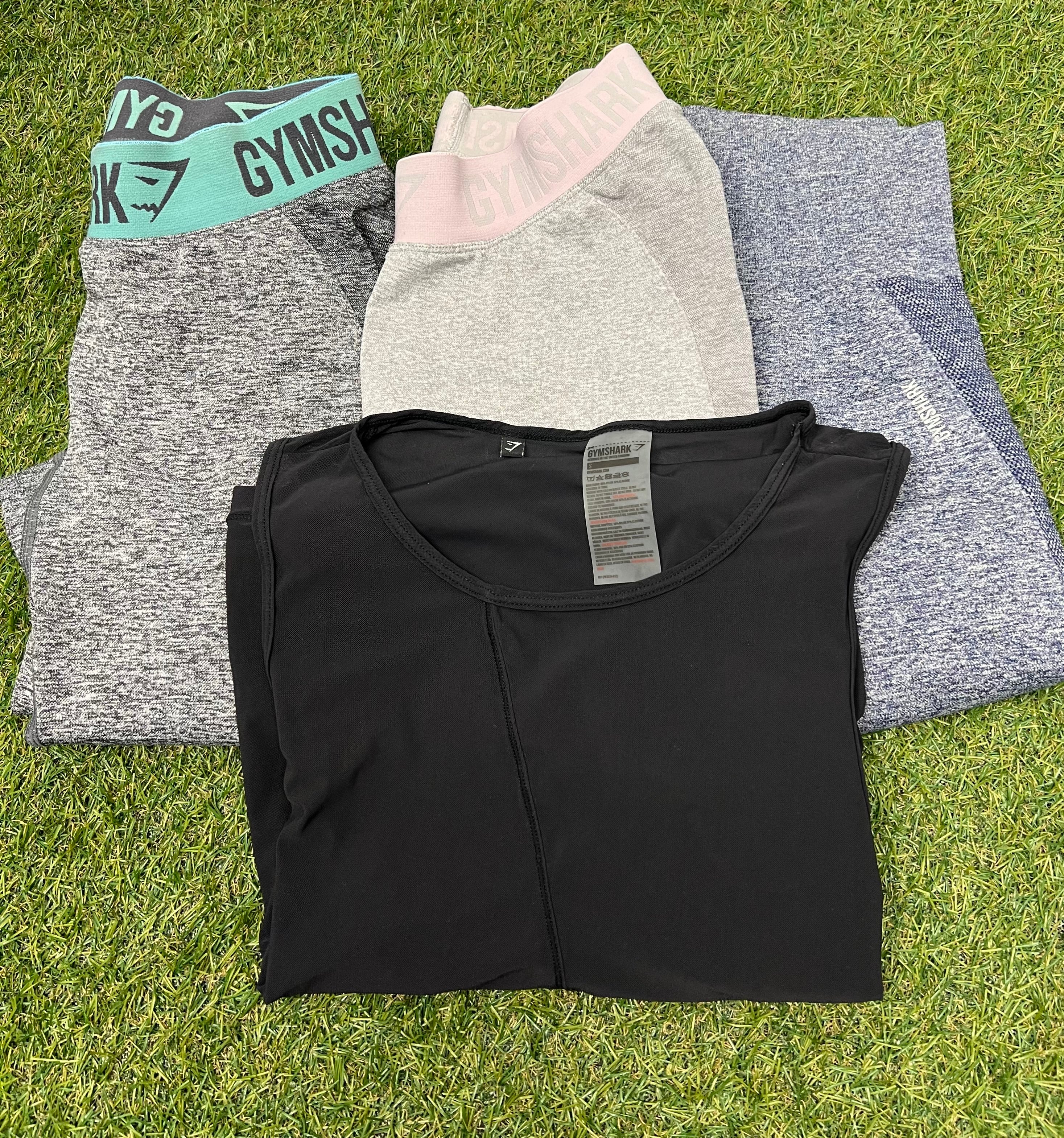 Folded Gymshark women's activewear set laid on grass including two pairs of seamless leggings in grey and blue heather, and a black fitted top