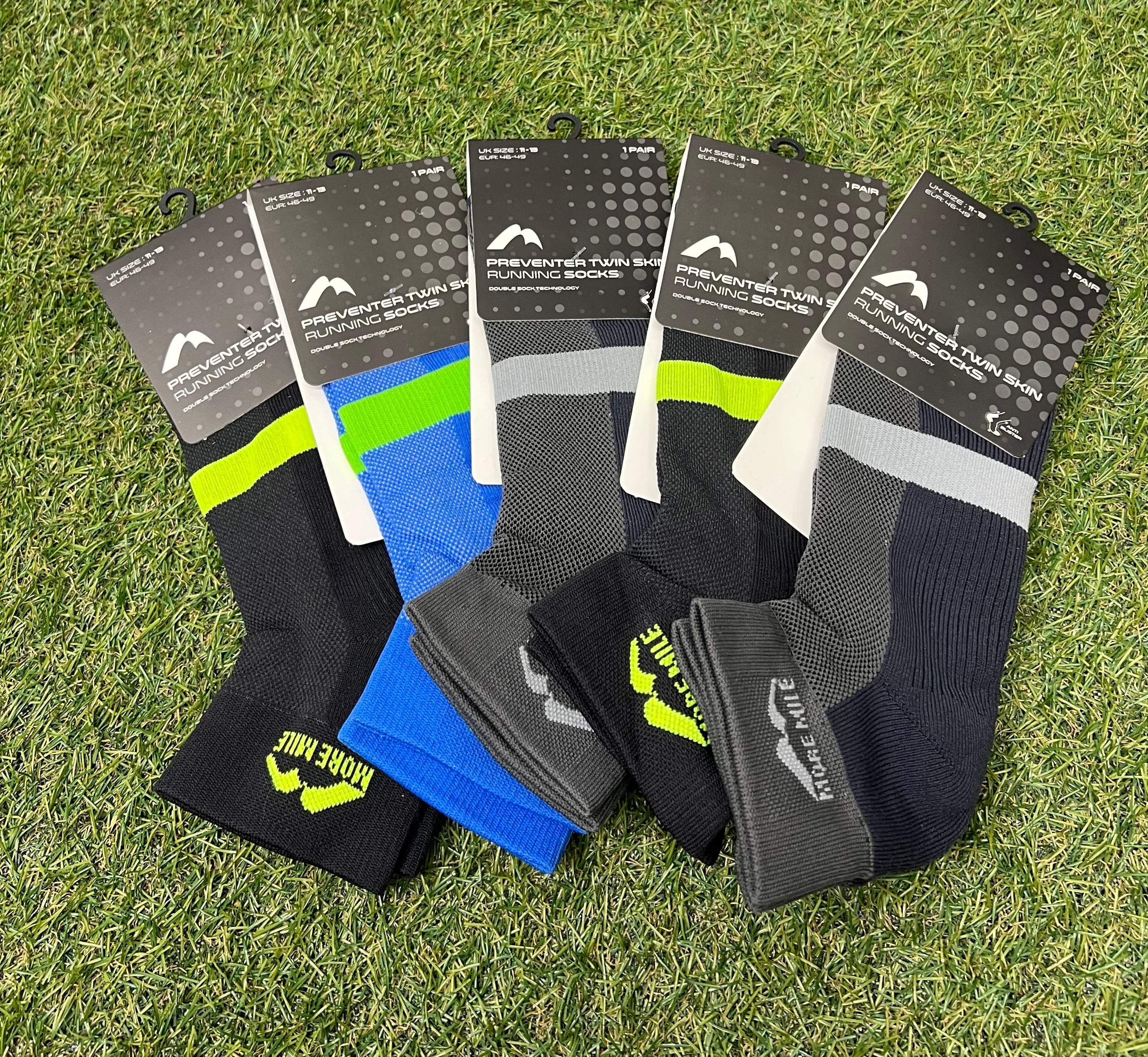Five pairs of More Mile Preventer Twin Skin Running Socks in assorted colours laid out on grass, showing front packaging