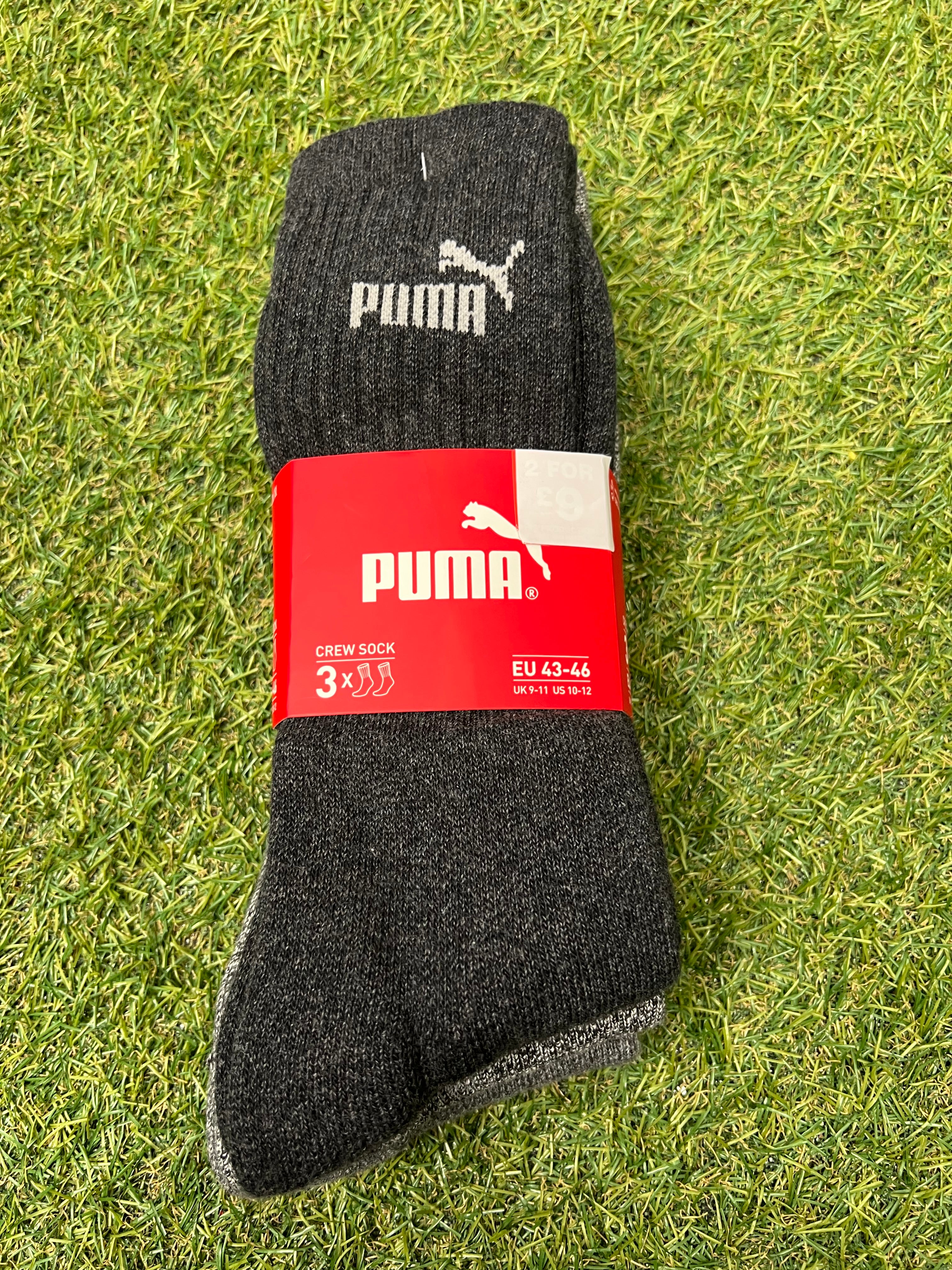 Pack of three dark grey Puma crew socks with red packaging on green grass background