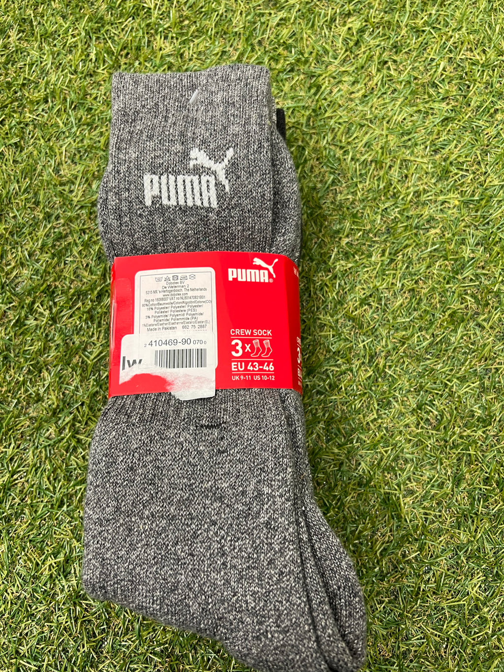 Pack of three light grey Puma crew socks with red packaging on green grass background
