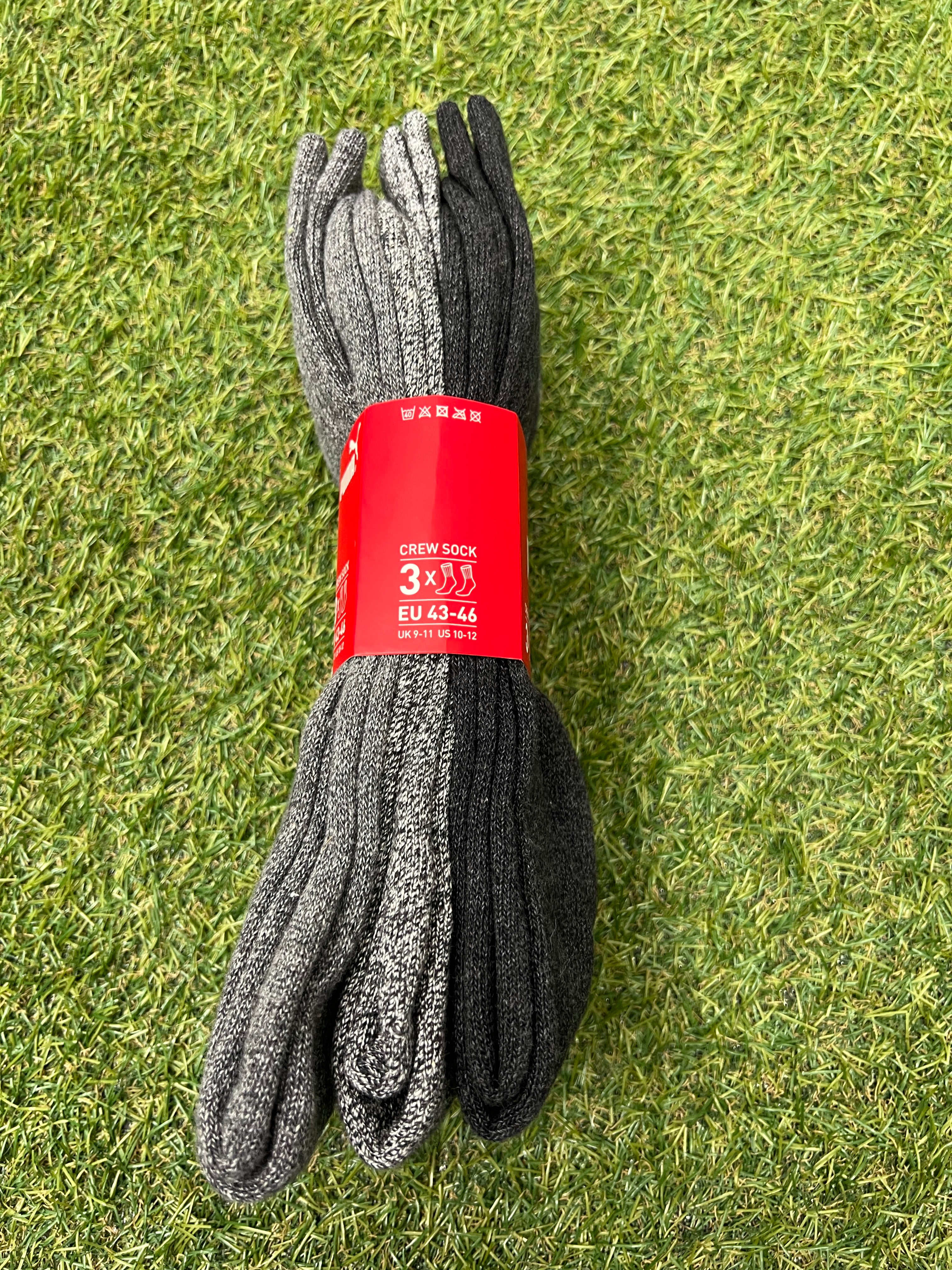 Pack of three Puma crew socks in black, dark grey and light grey arranged side by side with red packaging on green grass