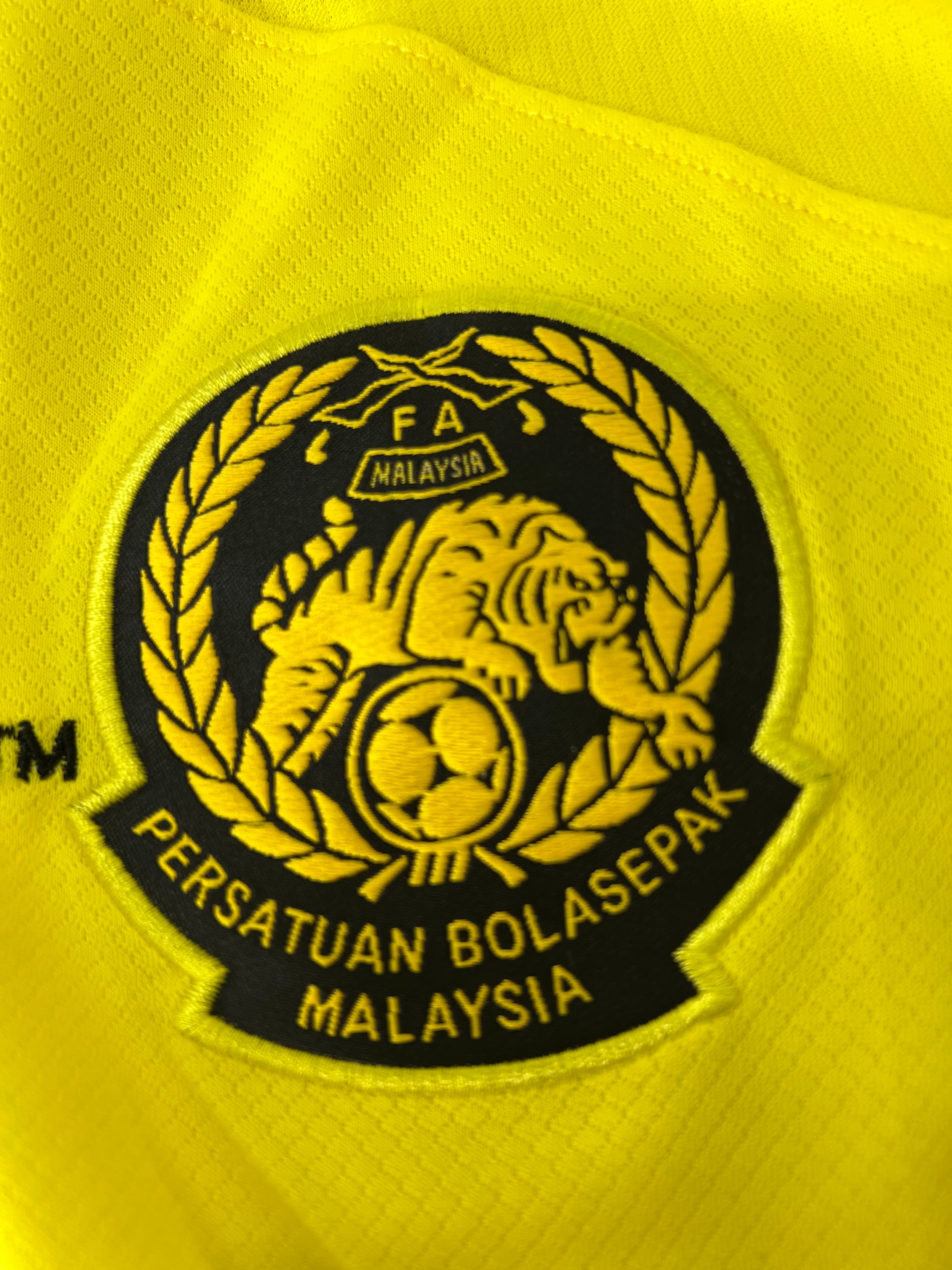 Close-up of embroidered Malaysia Football Association emblem on the chest of yellow football jersey
