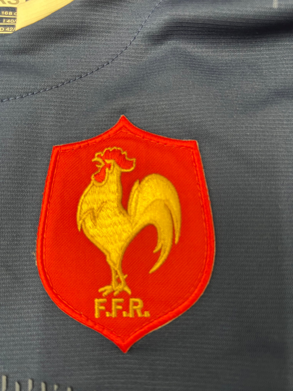 Close-up of the embroidered red and gold French Rugby Federation FFR rooster crest on jersey front