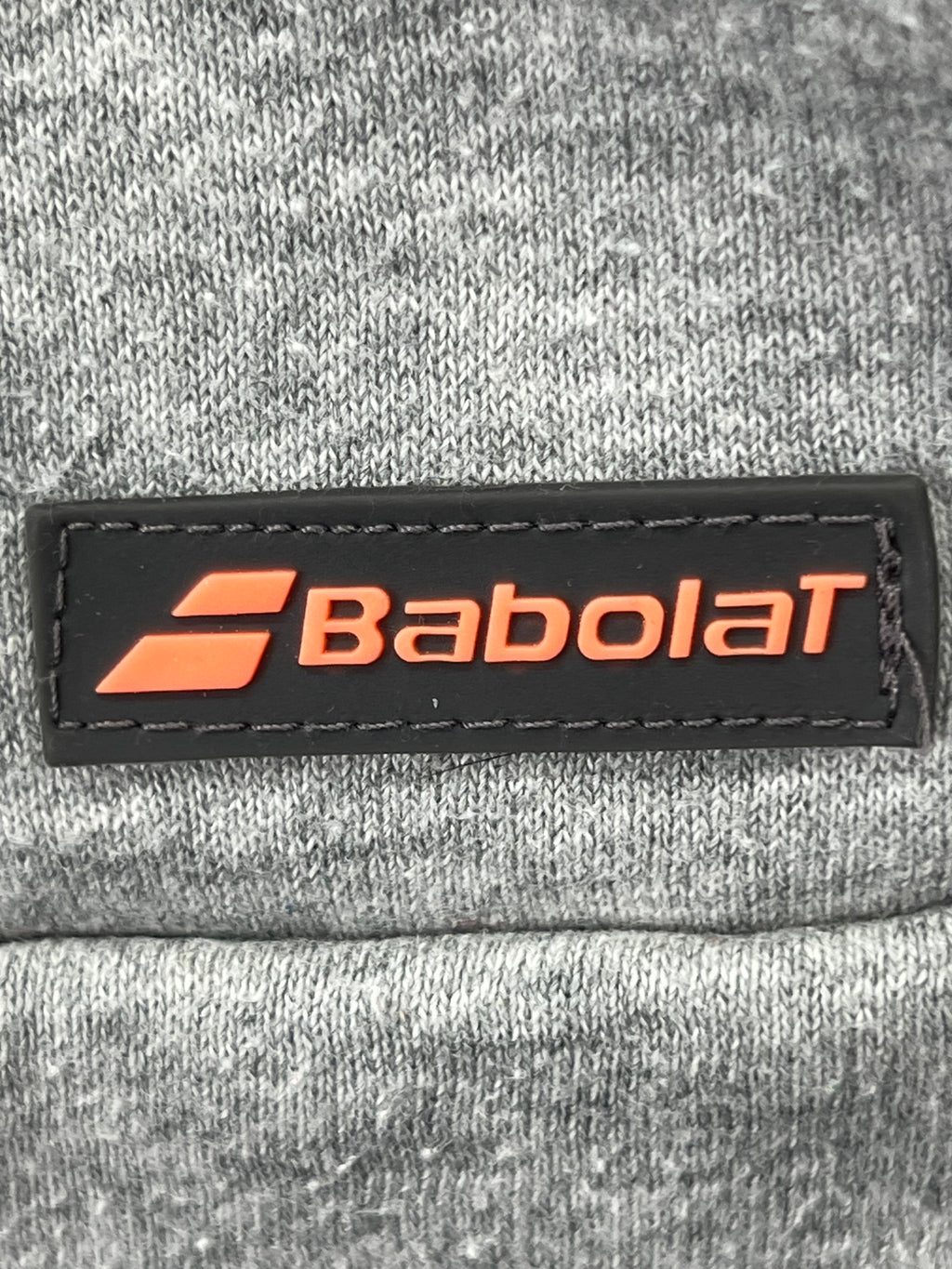 Close-up of black and orange Babolat logo patch sewn on grey hoodie fabric