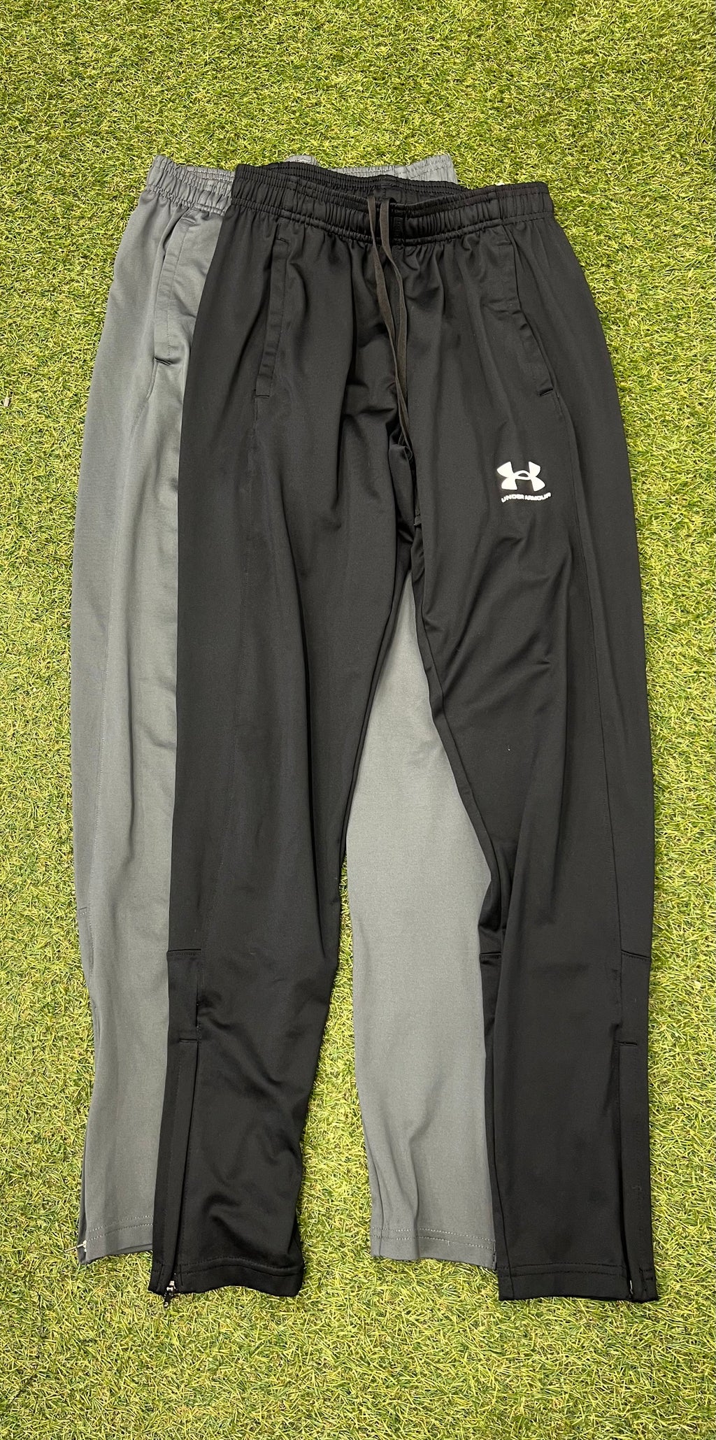 Two pairs of Under Armour tracksuit trousers in black and grey laid flat on grass