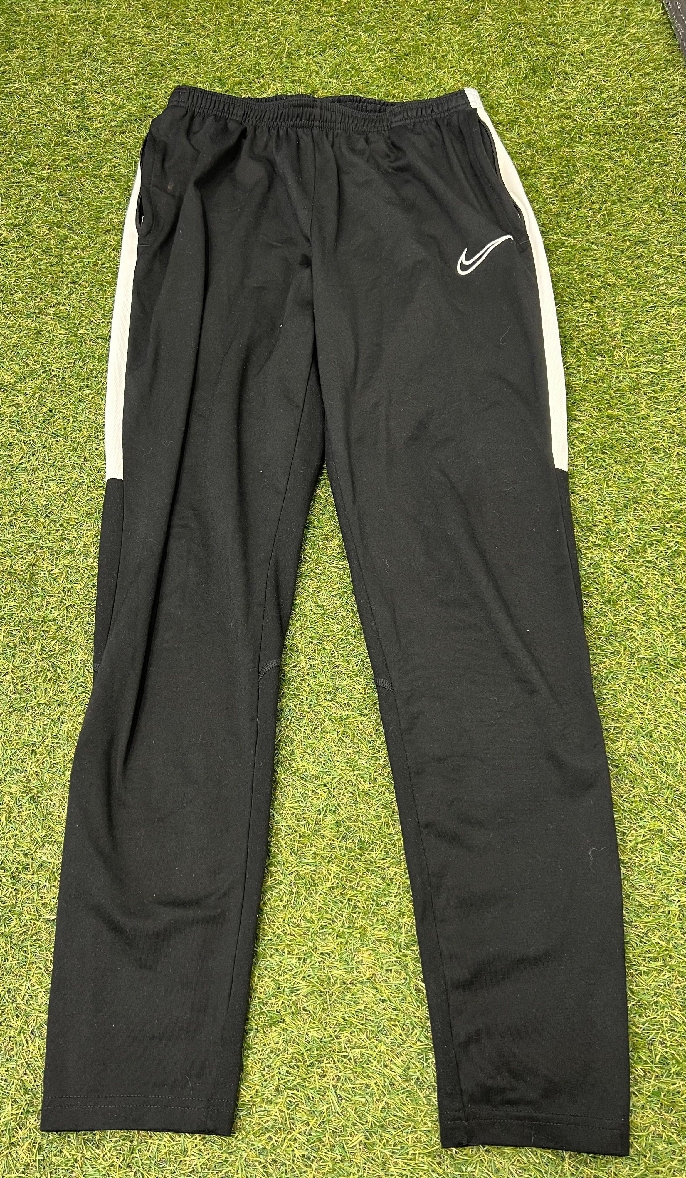 Front view of Nike black tracksuit bottoms with white side stripes on grass background
